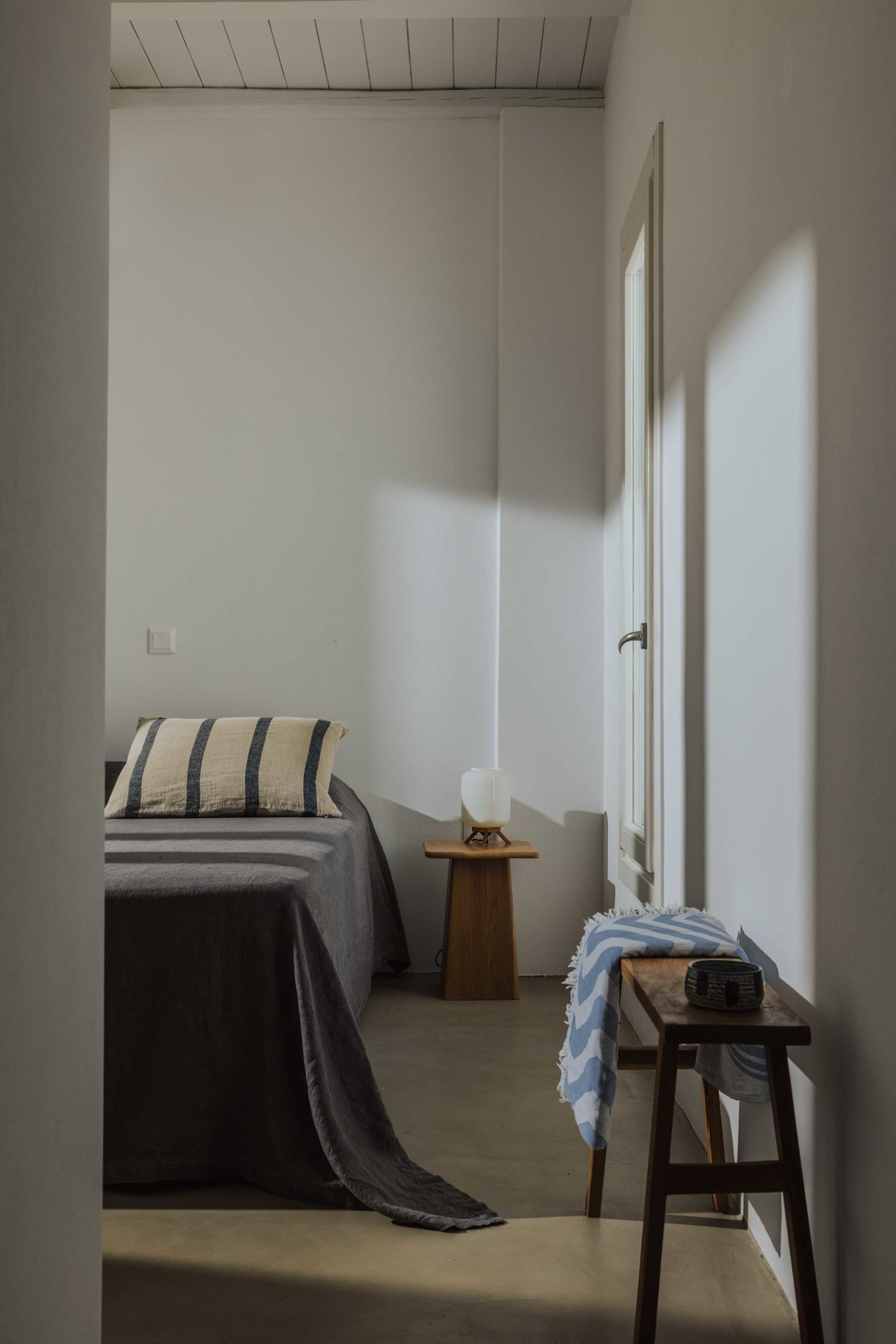 A minimalist bedroom with a concrete floor, a single nightstand with a lamp, and a striped pillow.
