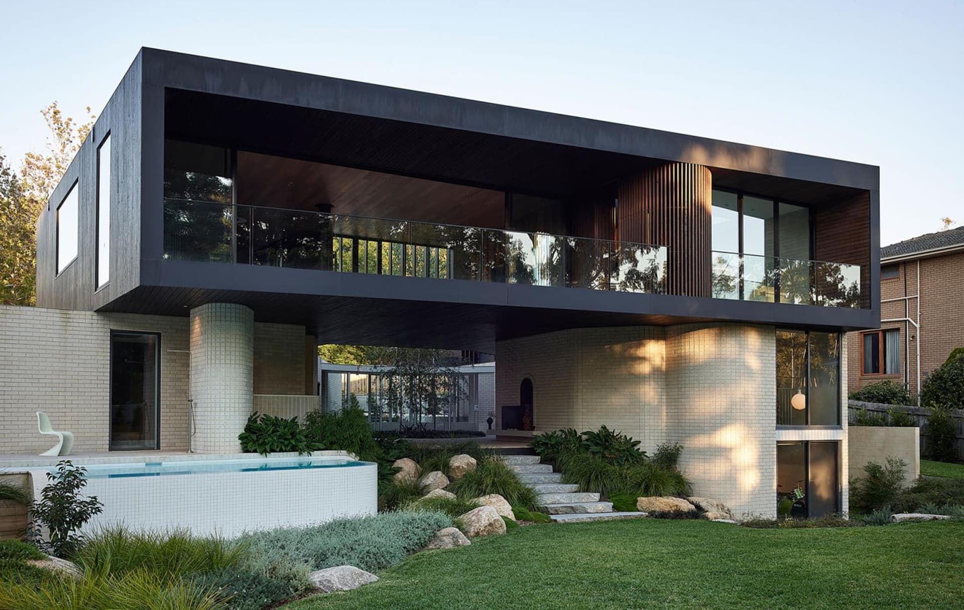 Modern residential exterior with brown brick cladding, concrete columns, and glass balustrades.