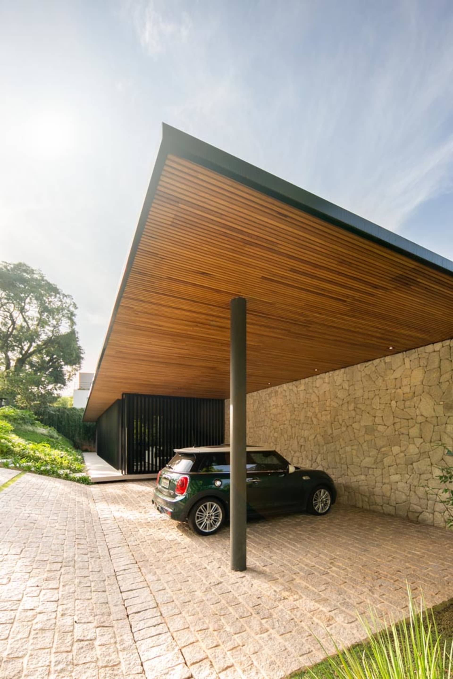 A modern carport with a wooden roof and stone walls featuring a green car parked underneath it on a sunny day.