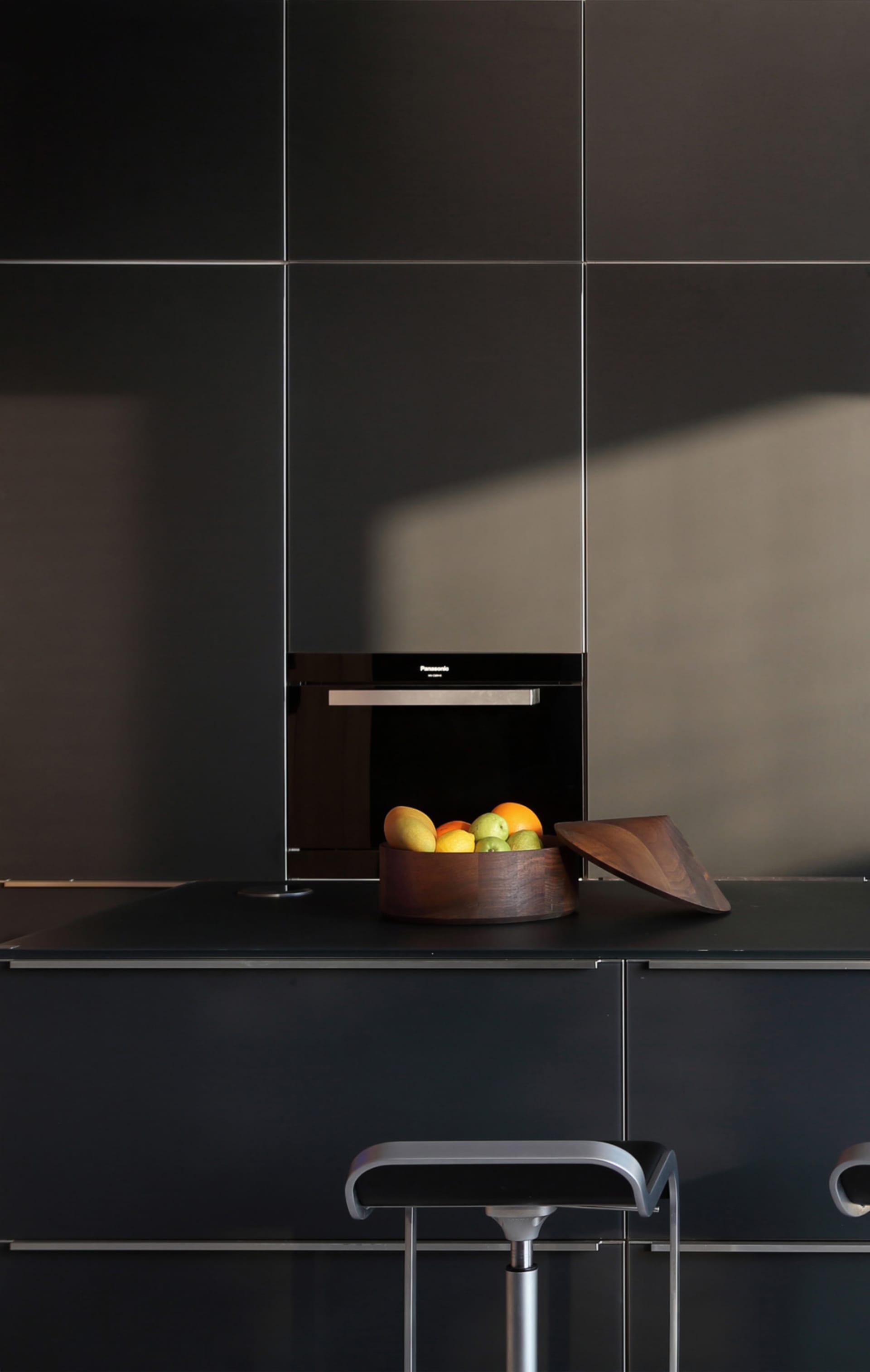 A modern kitchen interior featuring sleek, black cabinets and a countertop, with a wooden bowl containing a variety of fruits