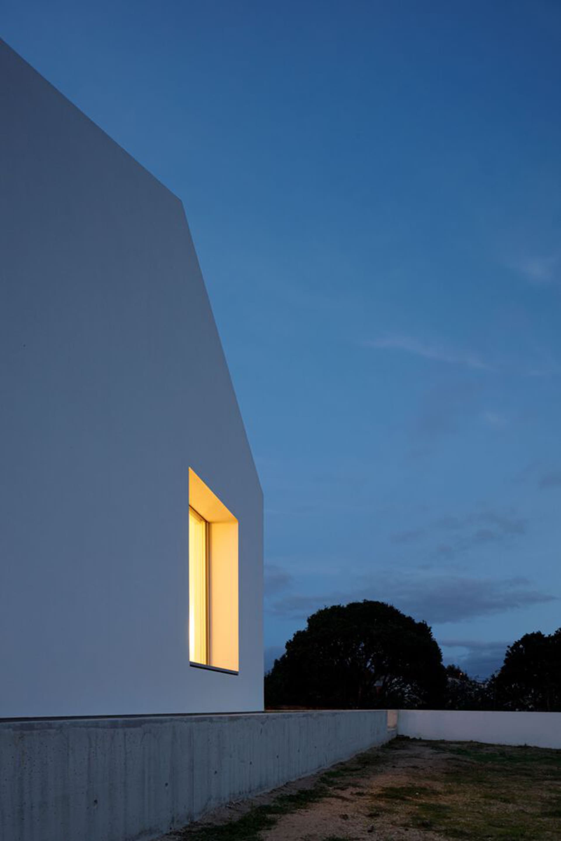A modern white house with geometric concrete cuts, seen at dusk with illuminated windows.