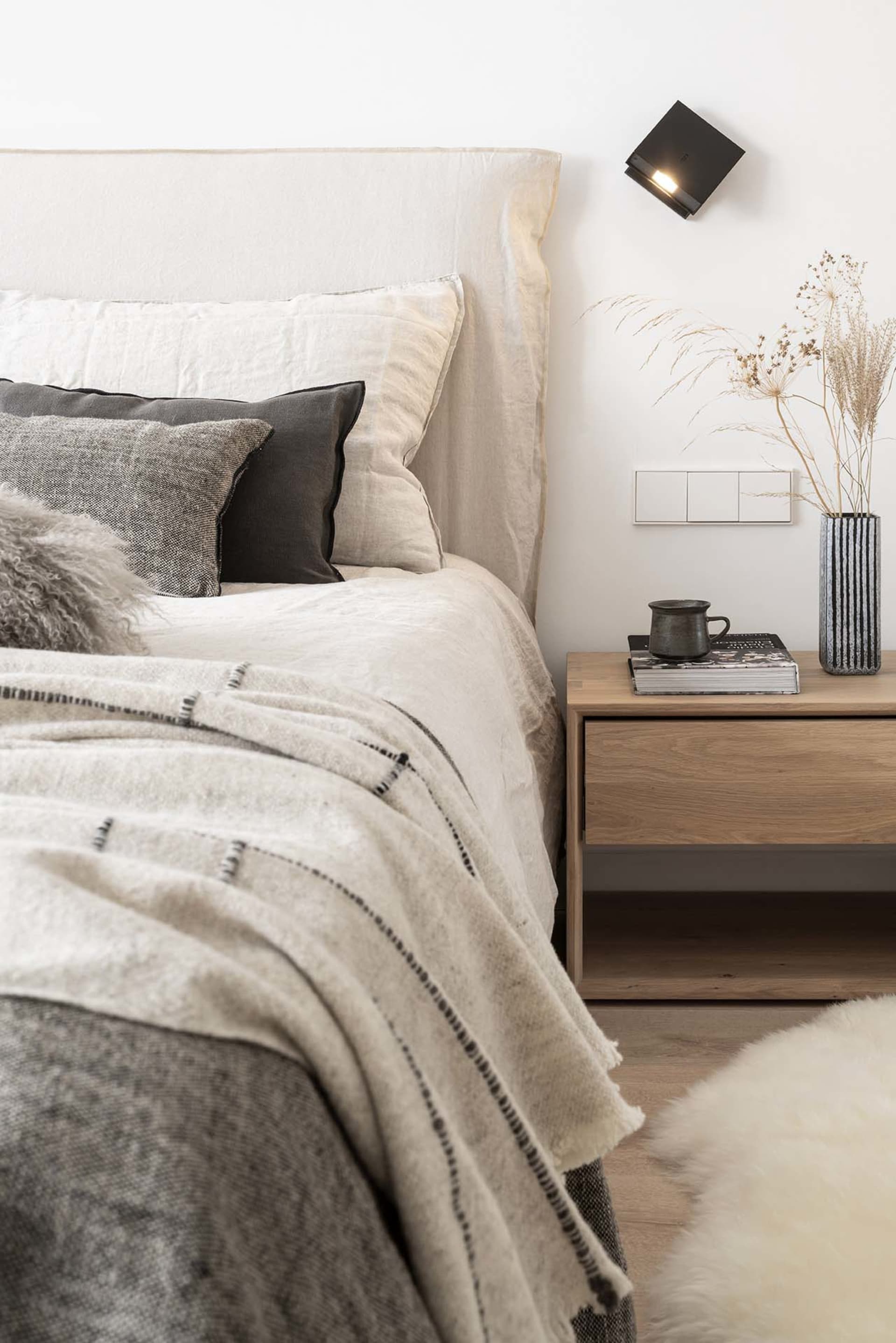 A modern minimalist bedroom with a bed featuring neutral-colored towels and a wooden nightstand with a vase of dried grasses 