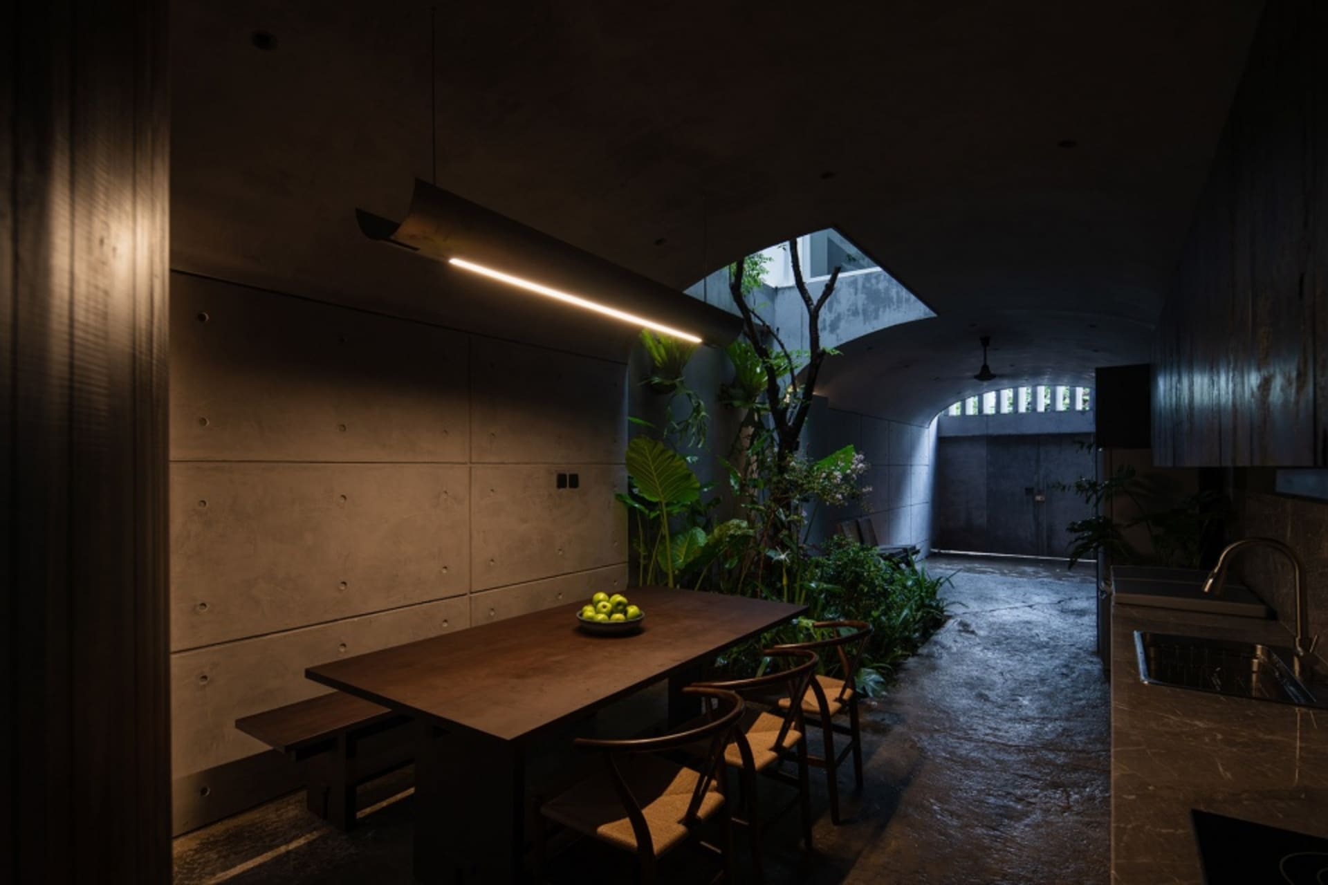 An industrial-themed dining room with a wooden table, chairs, and concrete walls, illuminated by a modern linear light fixtur