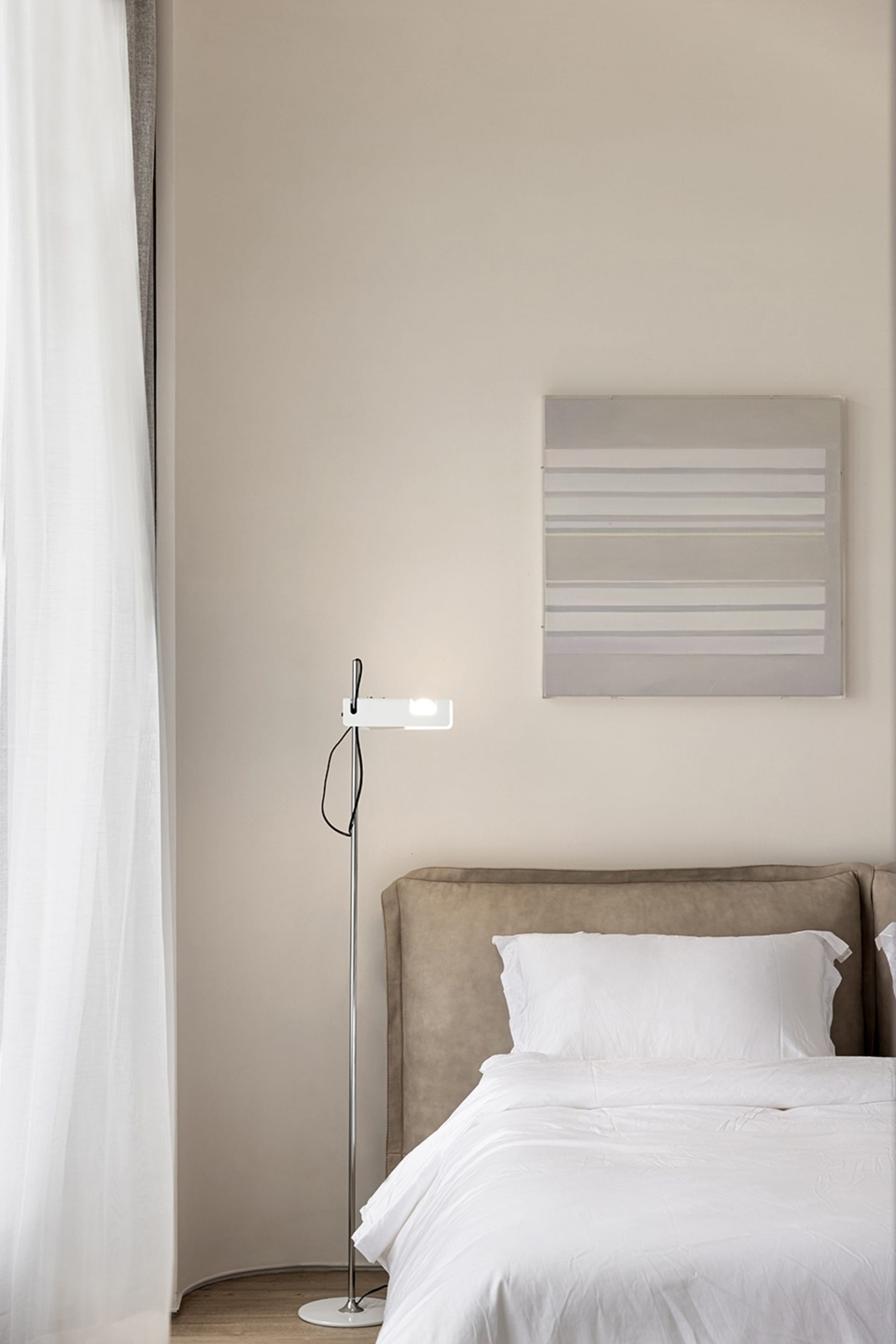 A modern bedroom featuring a white bed with a beige upholstered headboard, a minimalist floor lamp, and a light gray abstract