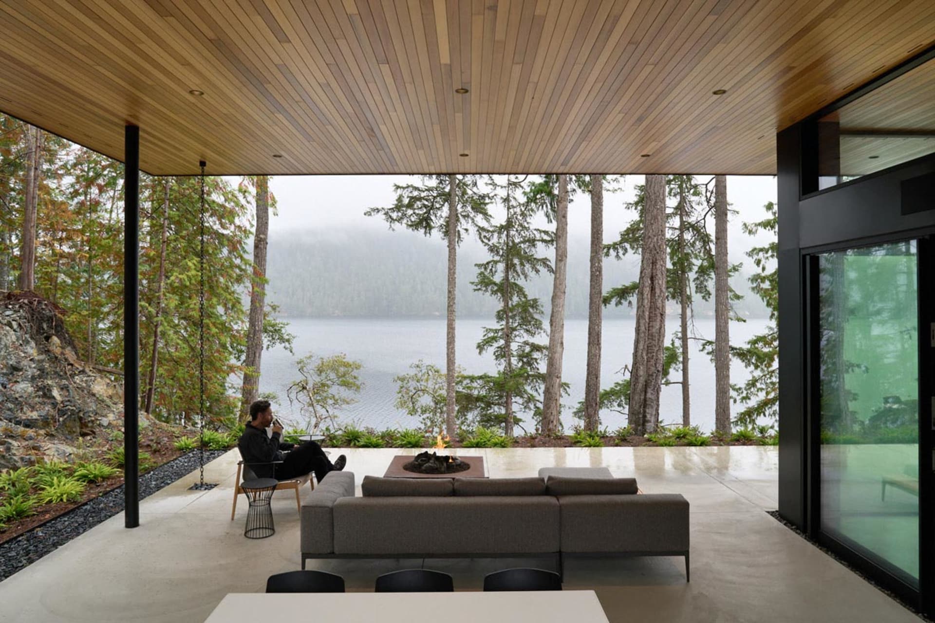 Modern living room with concrete furniture, glass walls, and a fireplace, overlooking a forest and lake.