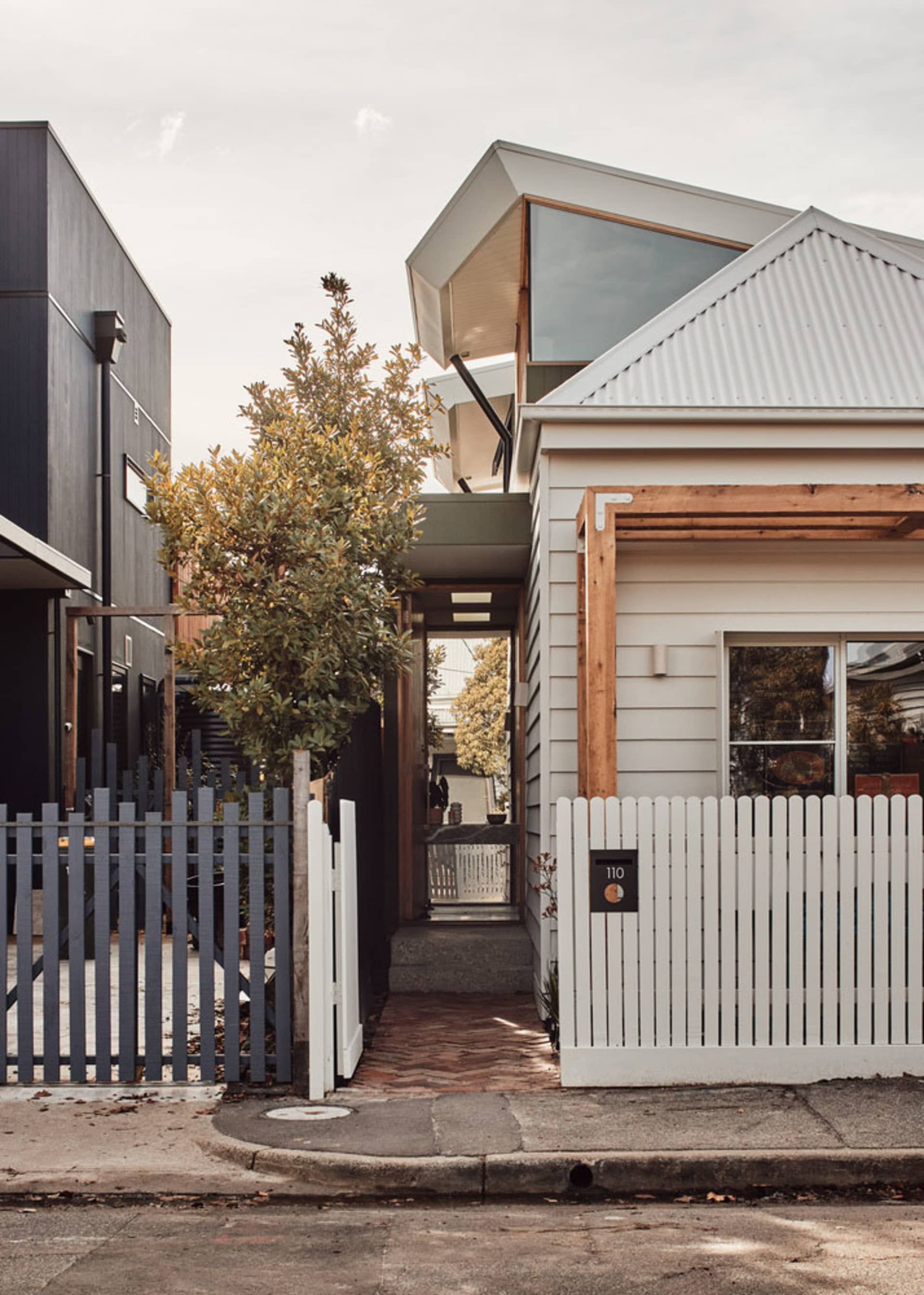 Modern house with wooden and metallic accents, featuring a white picket fence and a corrugated metal roof.