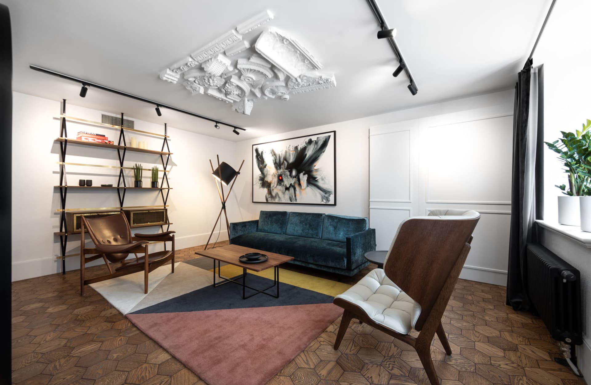 A modern minimalist living room featuring a blue velvet sofa, wooden chairs, and a geometric rug.