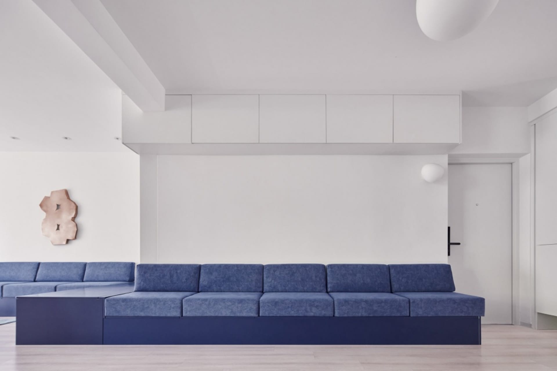 A modern minimalist living room featuring a blue sofa with a dark blue base and a rectangular metallic wall ornament.