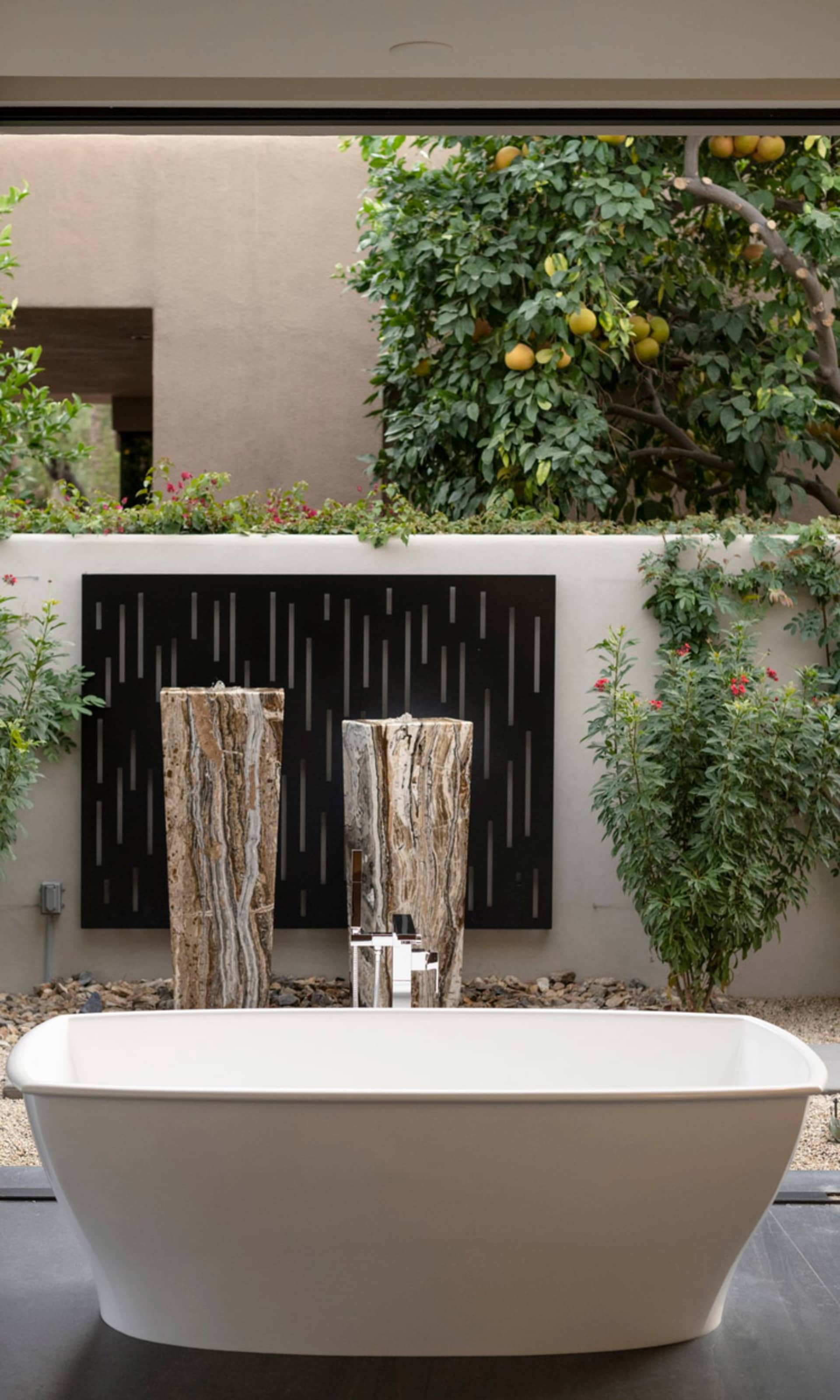 A minimalist bathroom with a white freestanding bathtub, marble columns, and a fruit-bearing tree in the background.