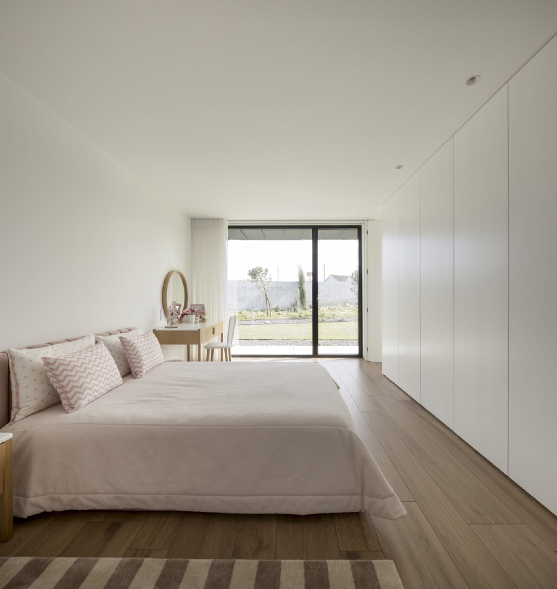 Modern minimalist bedroom with white walls, wooden flooring, and a large window overlooking a garden.