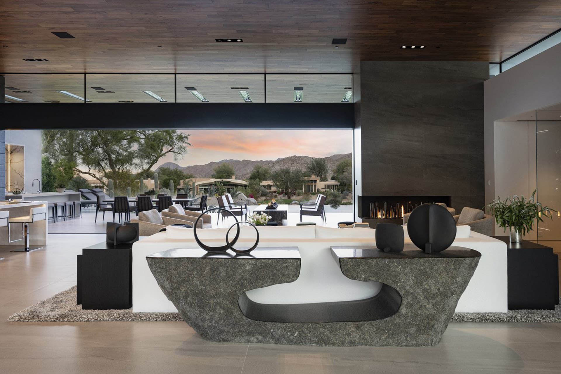 A modern living room with concrete furniture, wood accents, and a large glass window overlooking a desert landscape.