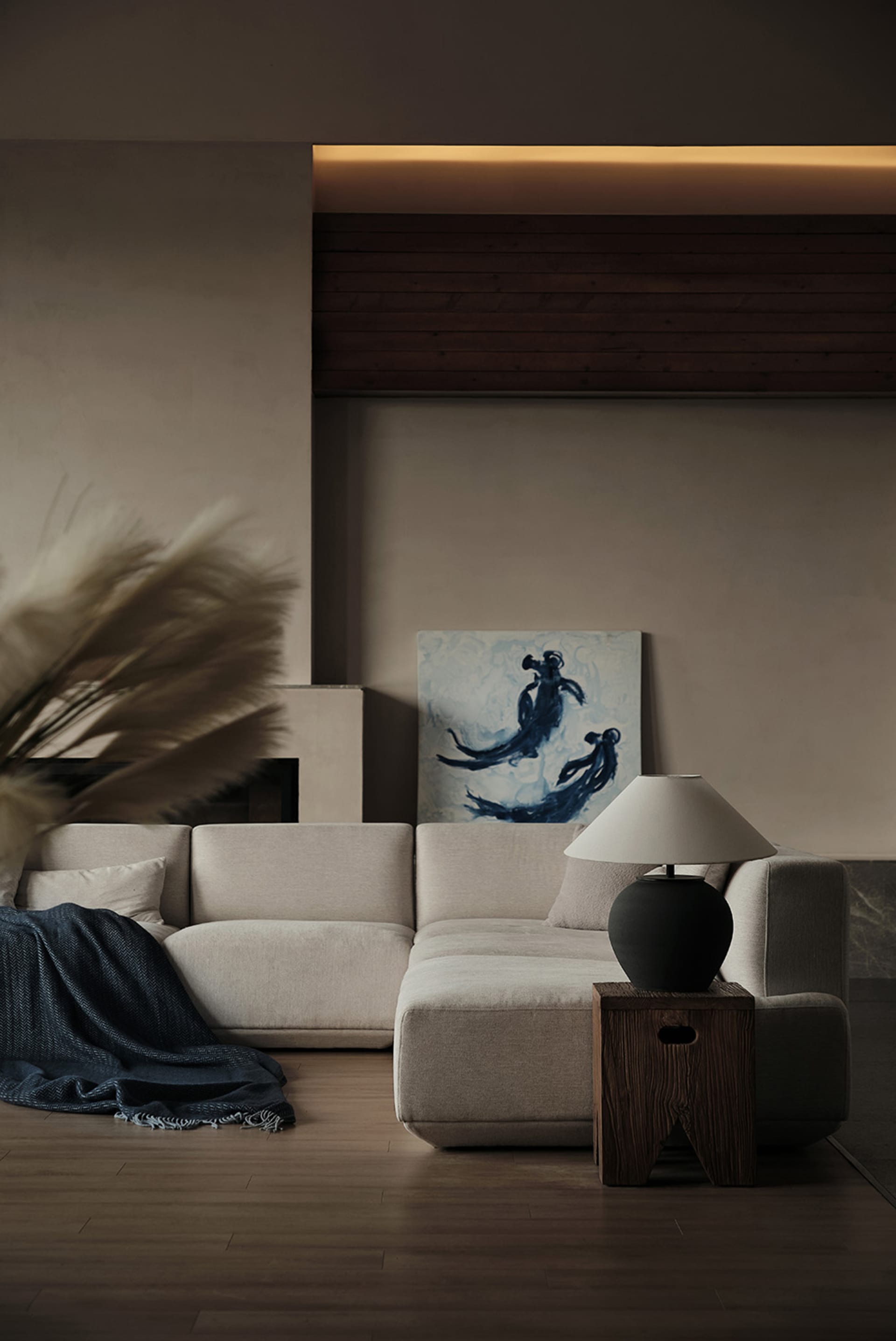 A modern living room with a beige sectional sofa, a dark lamp on a wooden side table, and a painting of silhouetted figures.
