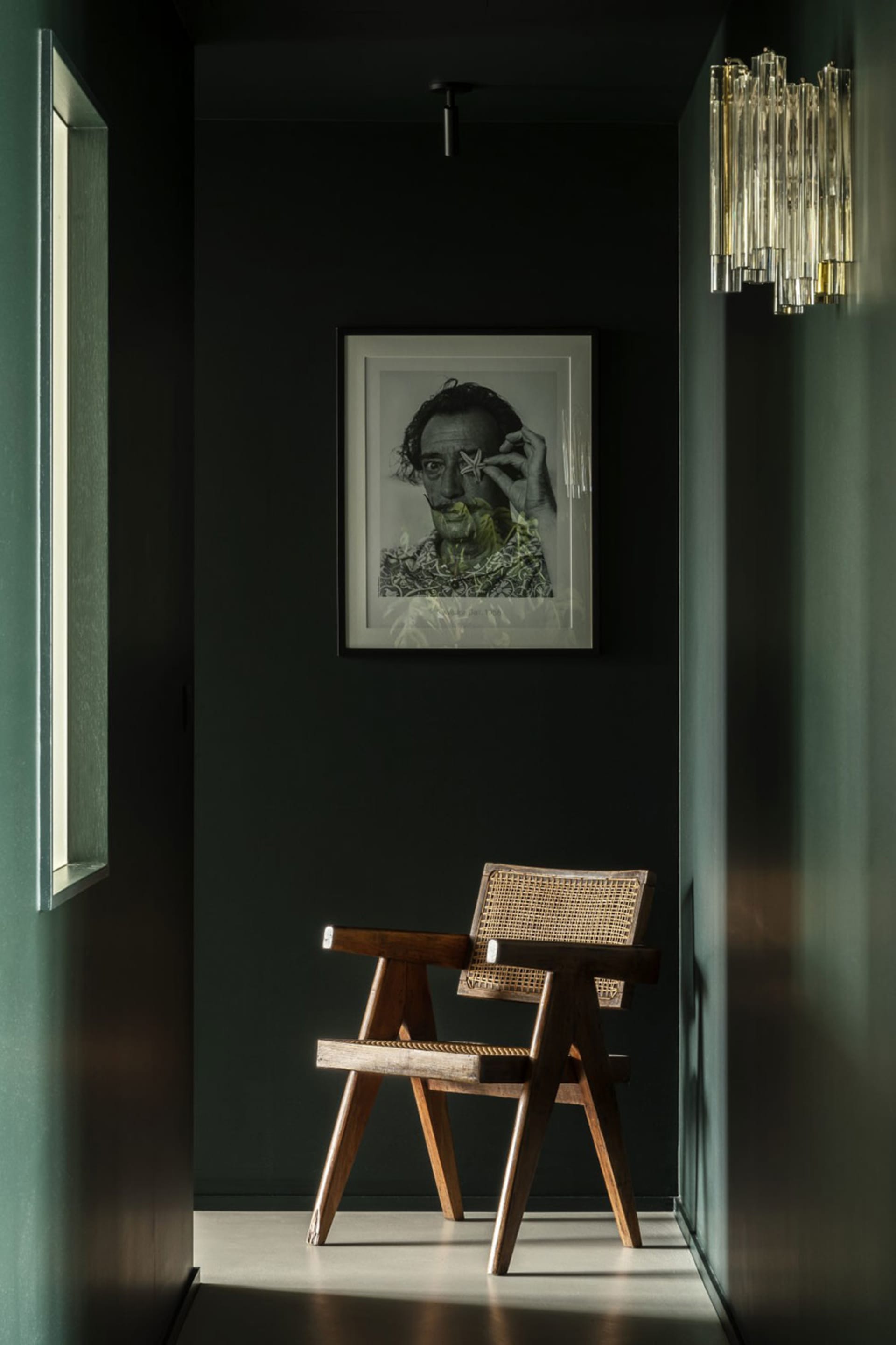A minimalist interior design featuring a wooden chair and a black and white portrait hanging on a dark green wall.