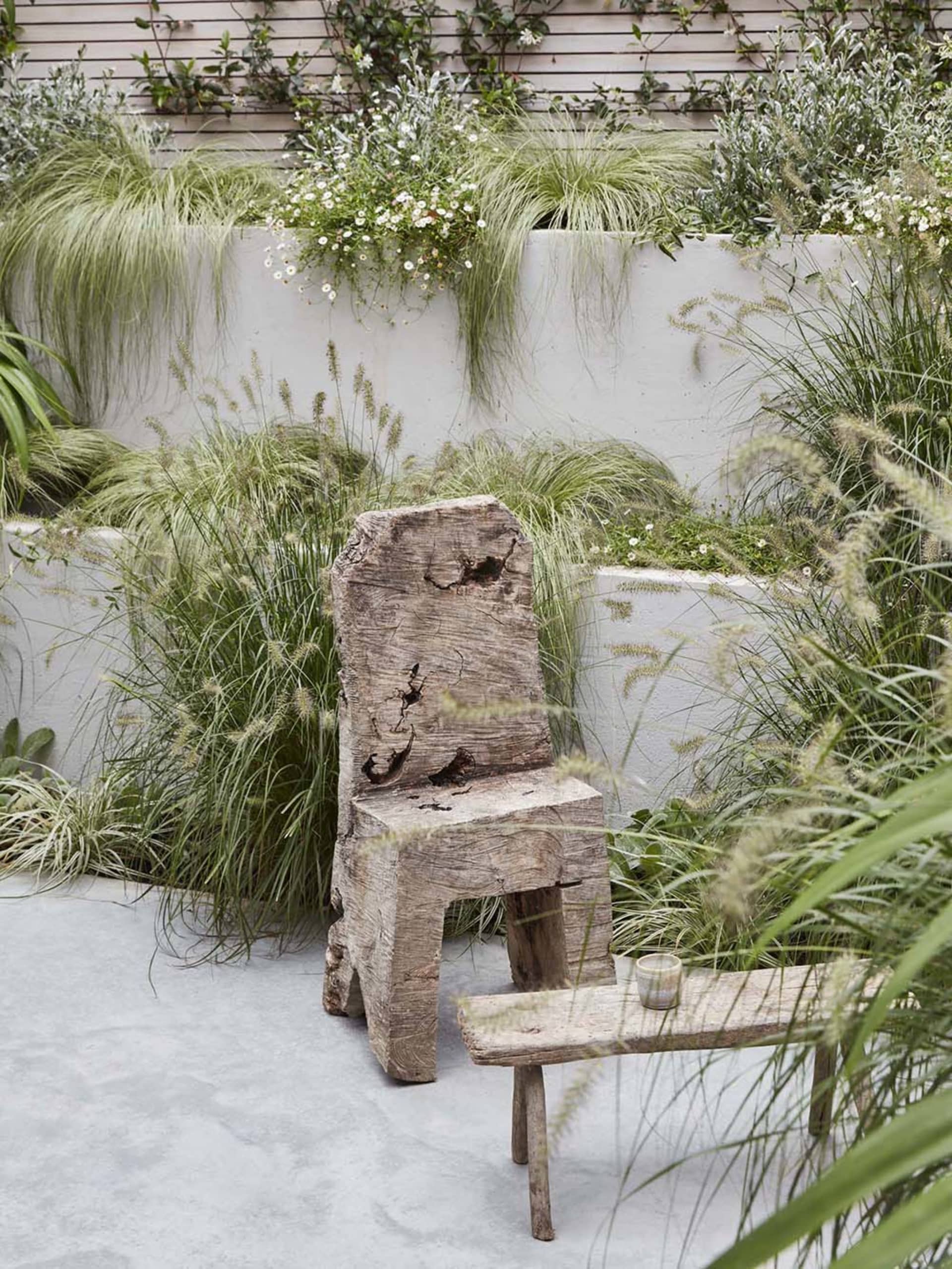 A rustic wooden chair and table in an outdoor garden setting surrounded by green foliage and plants.