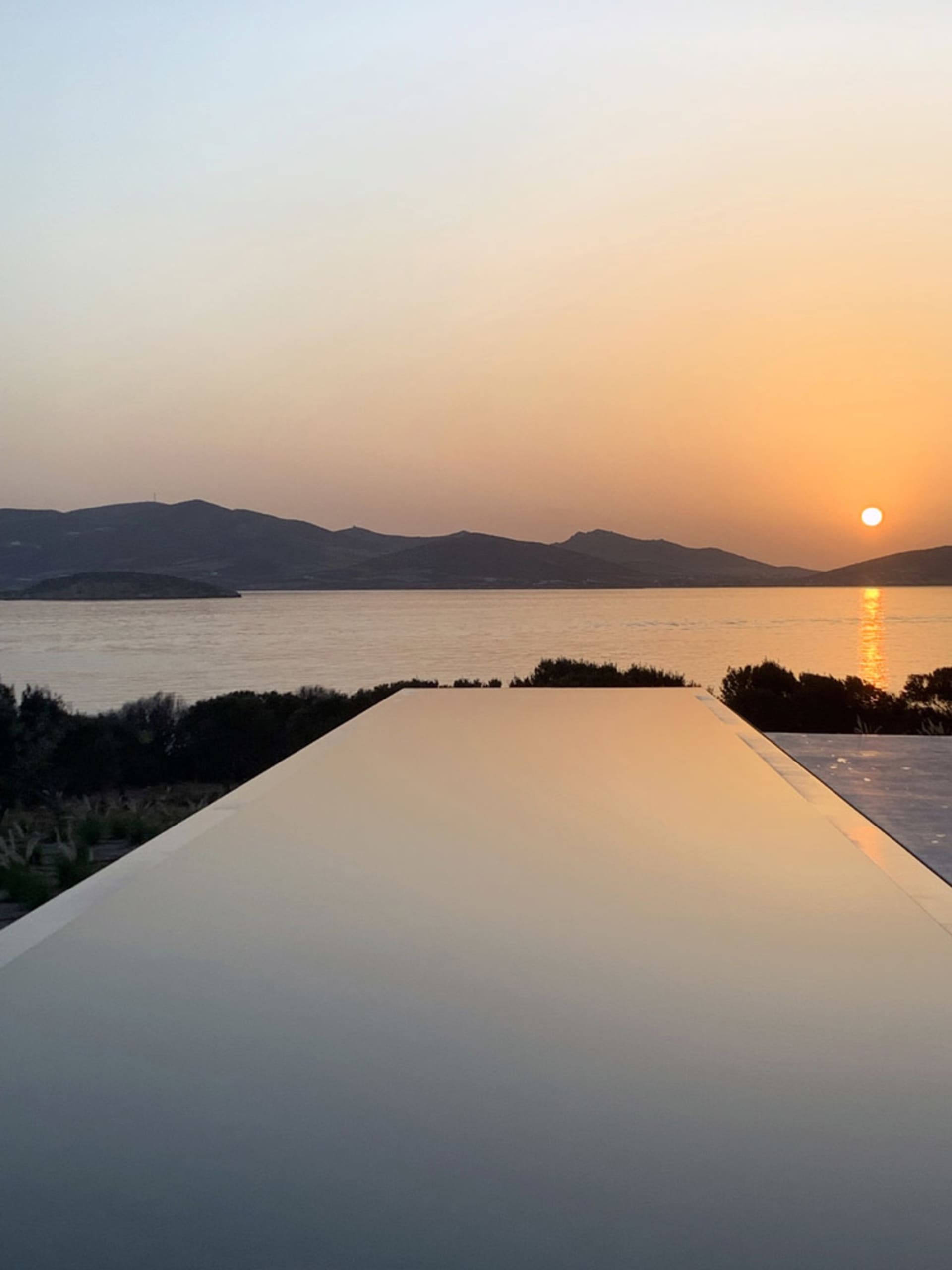 A minimalist swimming pool with an infinity edge overlooks a sunset view over the sea and mountains.