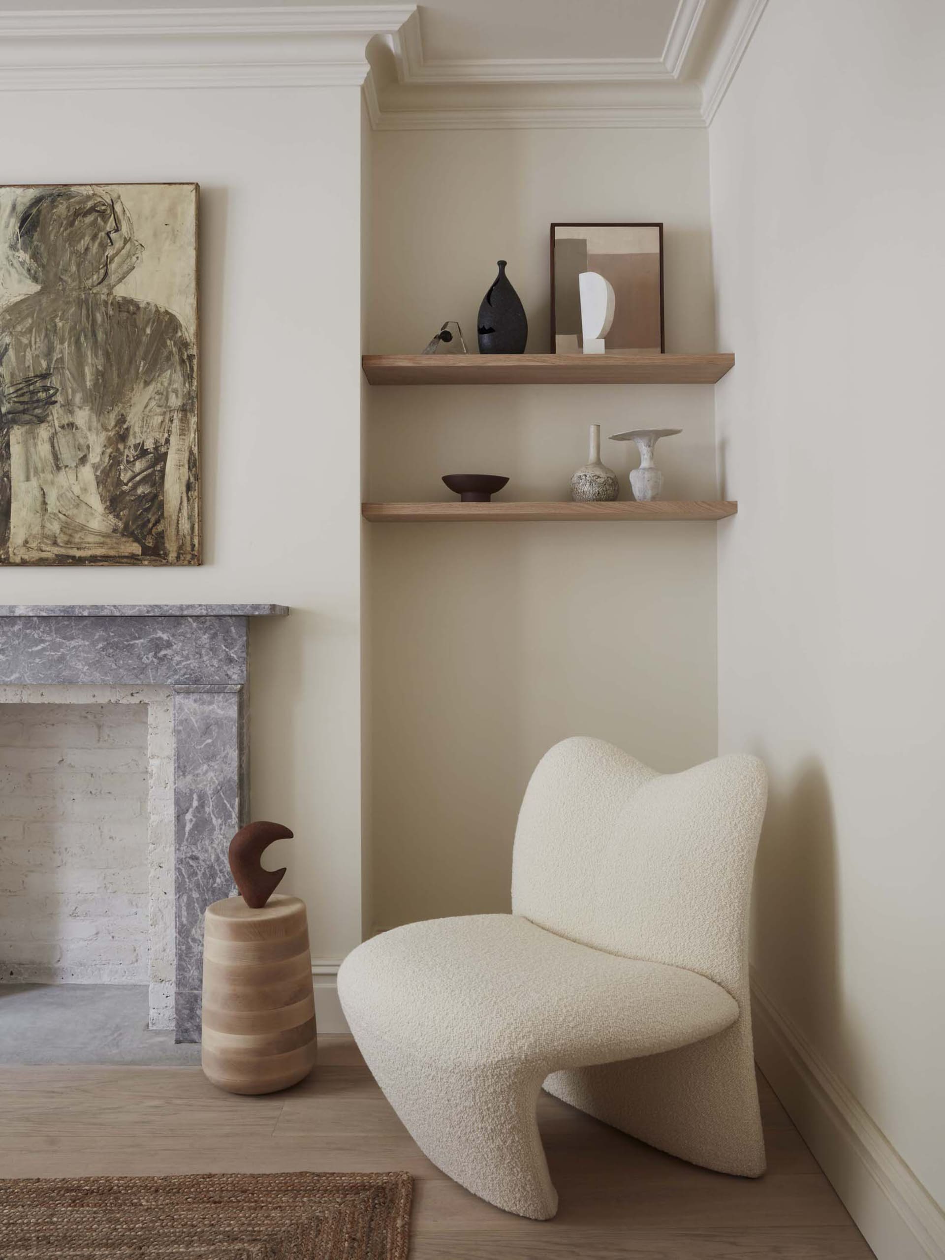 A cozy corner of a minimalist residential interior featuring a white textured lounge chair, a marble fireplace, wood shelves,