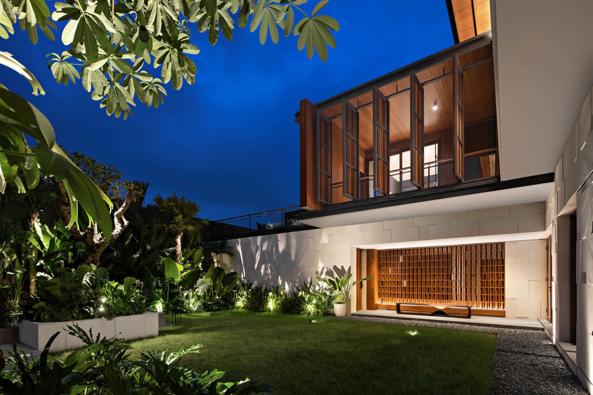 Night view of a minimalist residential garden with concrete walls, wooden louvers, and lush greenery.