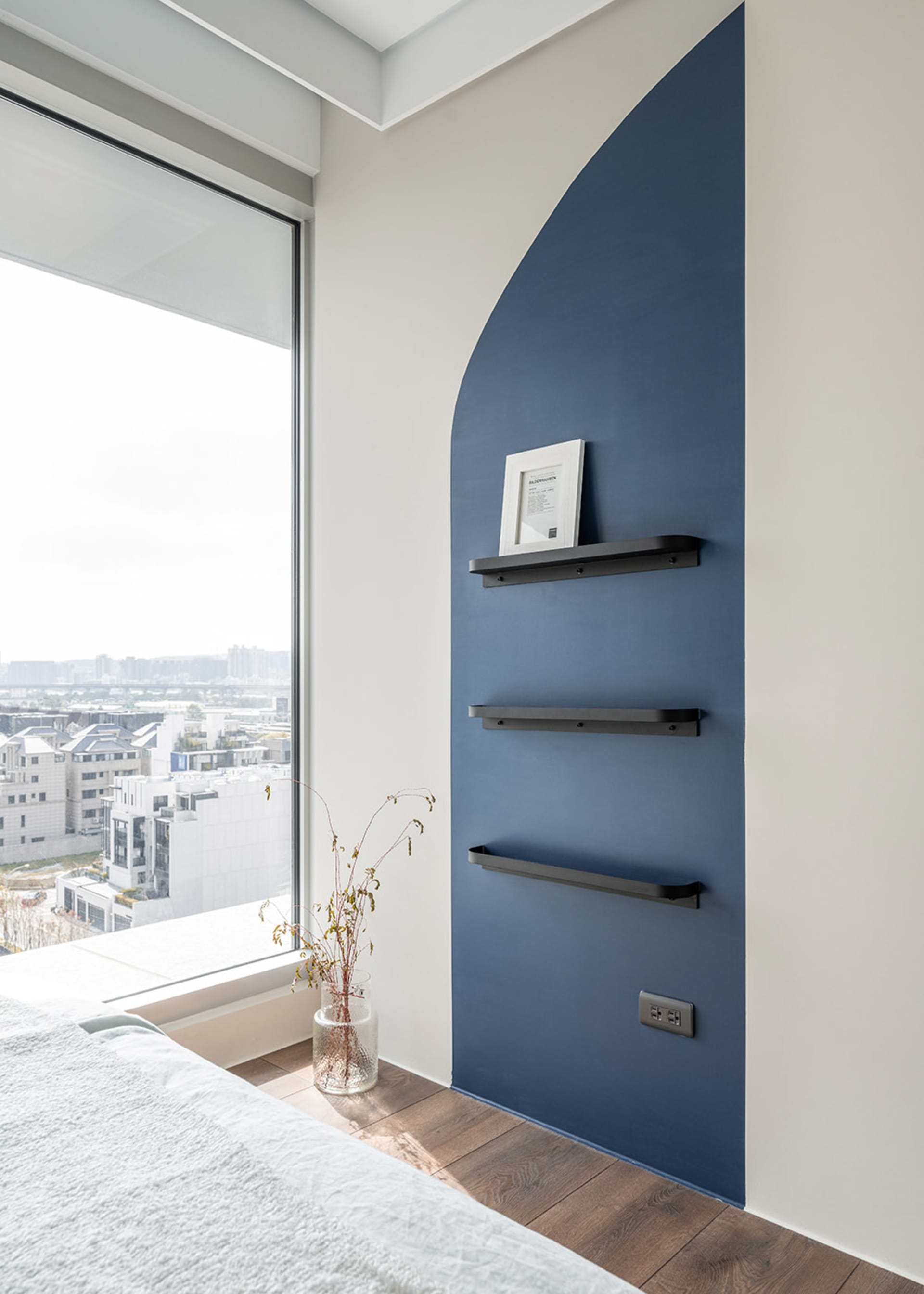 A modern bedroom featuring a blue accent wall with sleek, black metal shelves and a large window overlooking a cityscape.