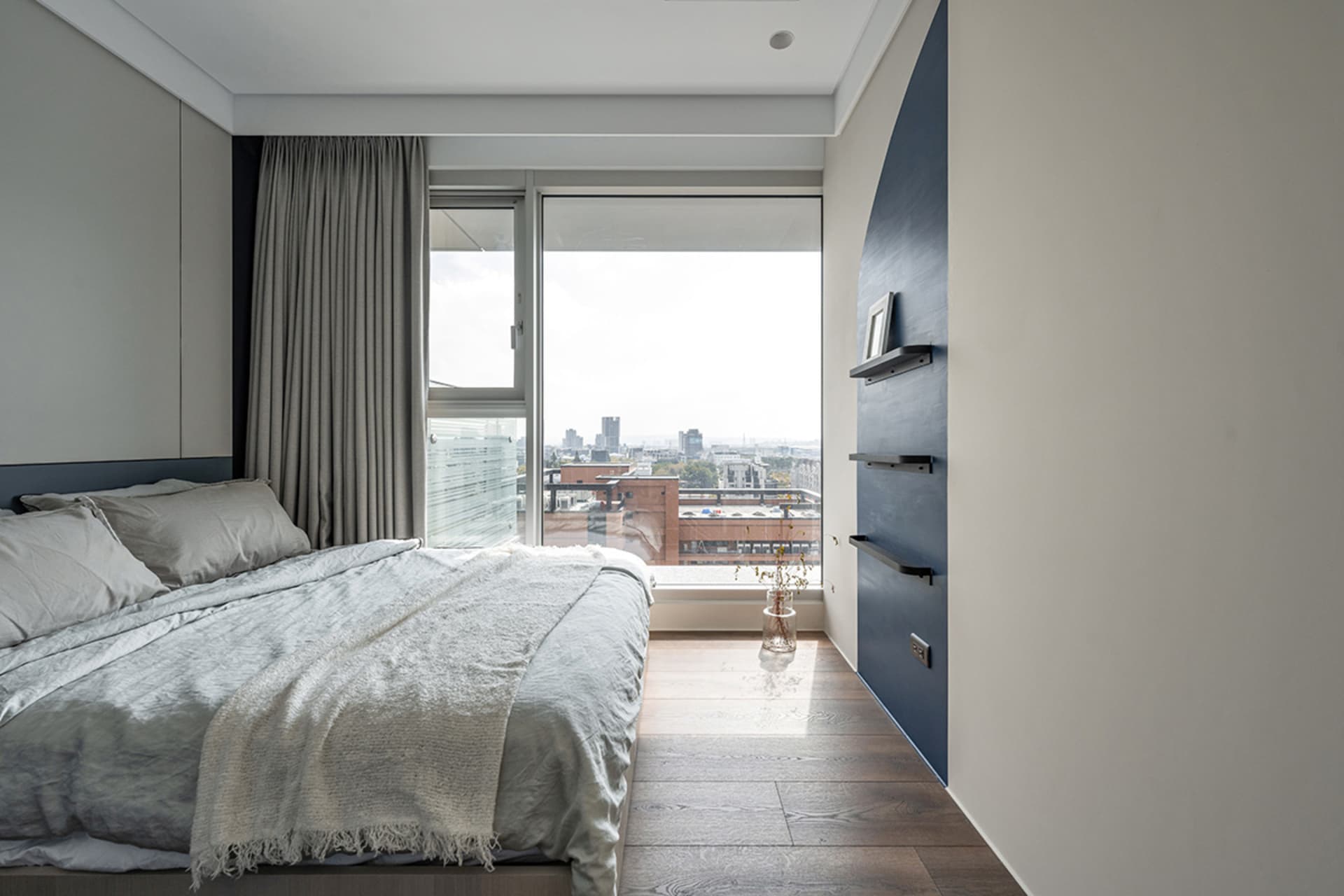A modern, minimalist bedroom with timber flooring, a large window overlooking a cityscape, and simple furniture.