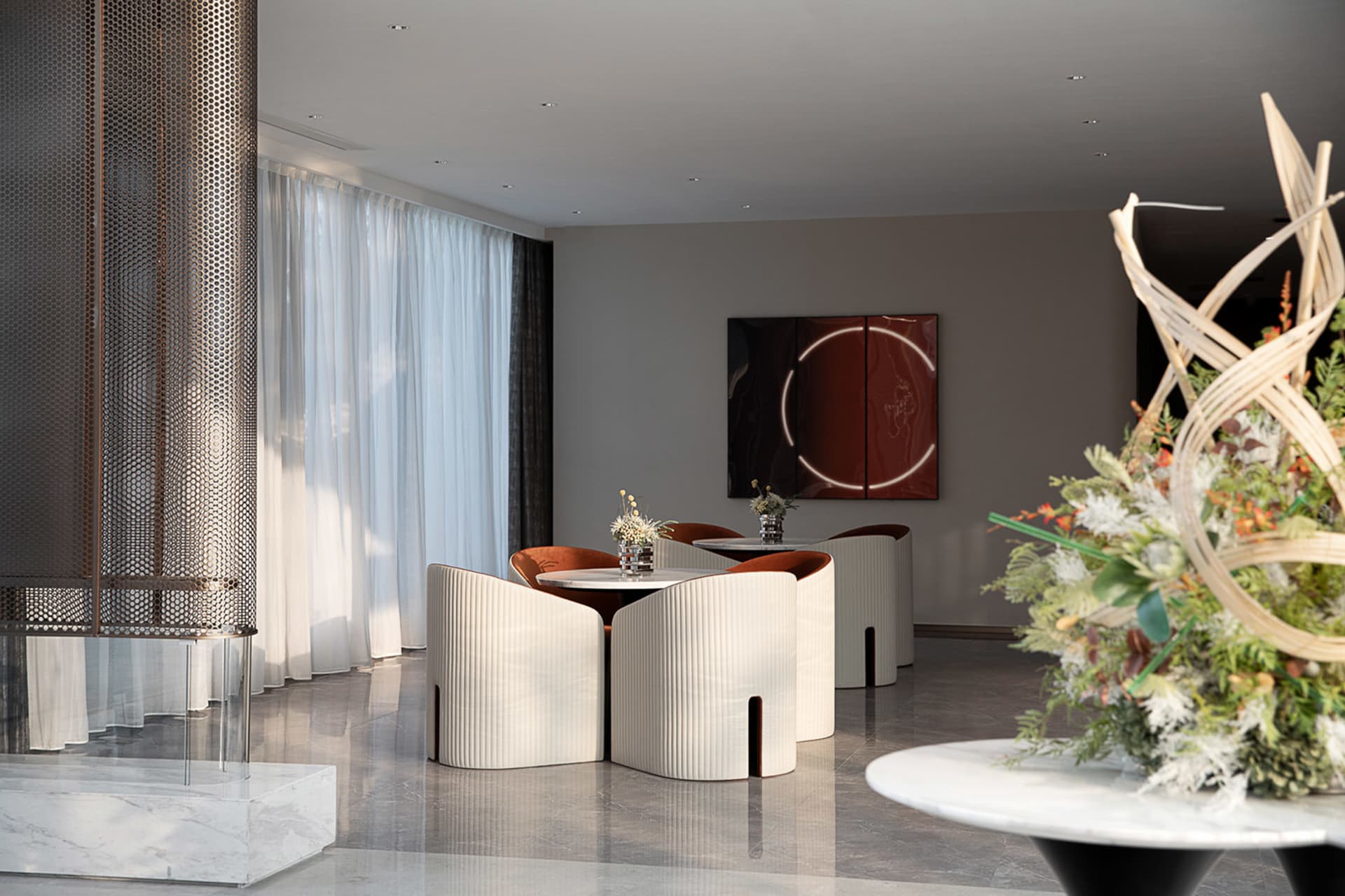 Modern hotel lobby featuring cream-colored plastic chairs and tables, with a large floral arrangement and a minimalist art pi