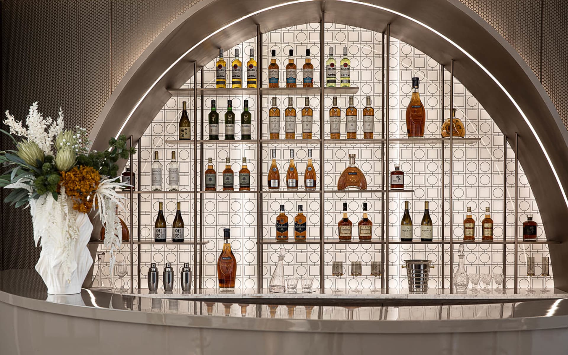 A modern metallic bar with a large arrangement of liquor bottles on a tiled wall.