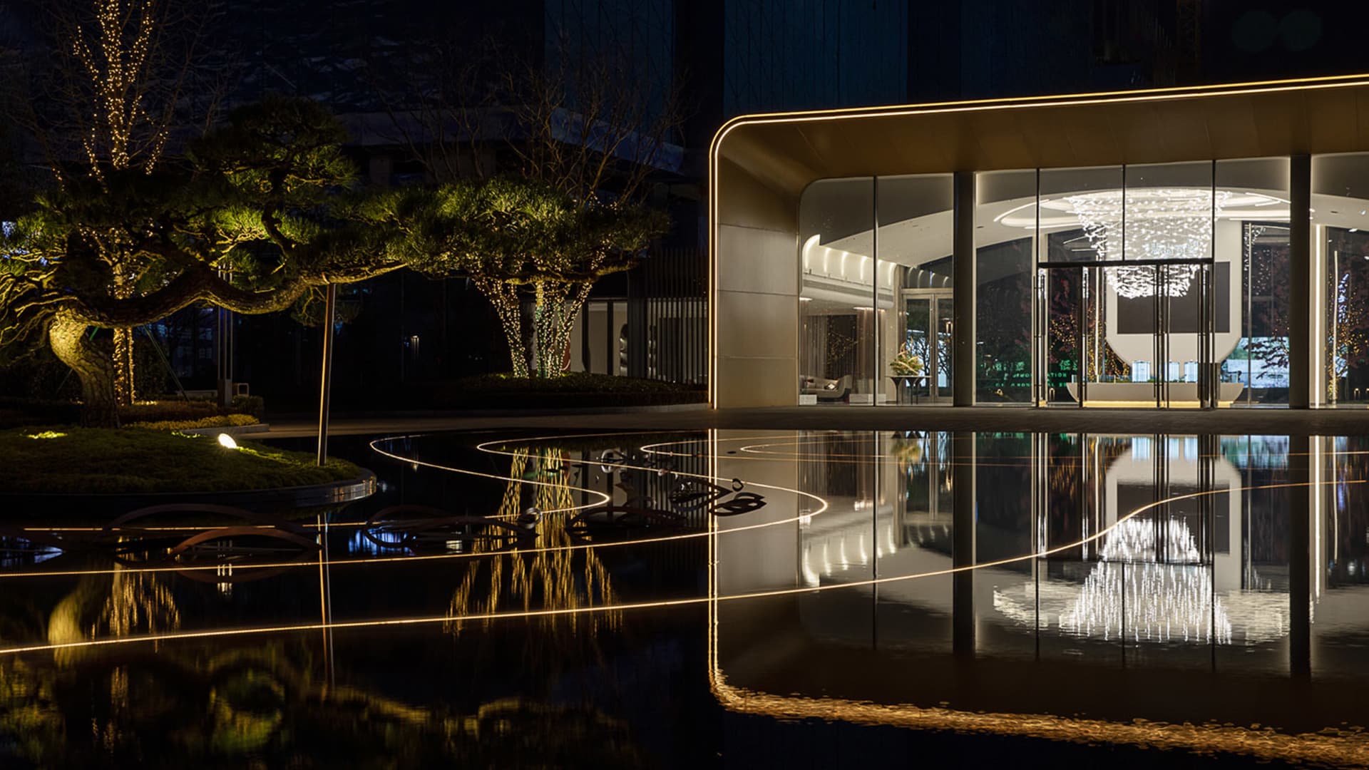 A modern entrance with glass doors, illuminated by warm lighting, reflecting off a water feature.