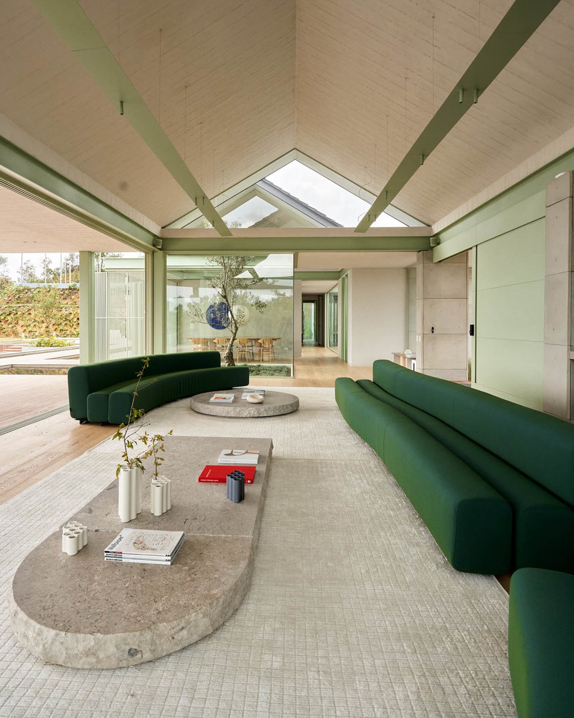 A modern minimalist interior with green sofas, concrete tables, and a high, wooden-beamed ceiling with skylights.