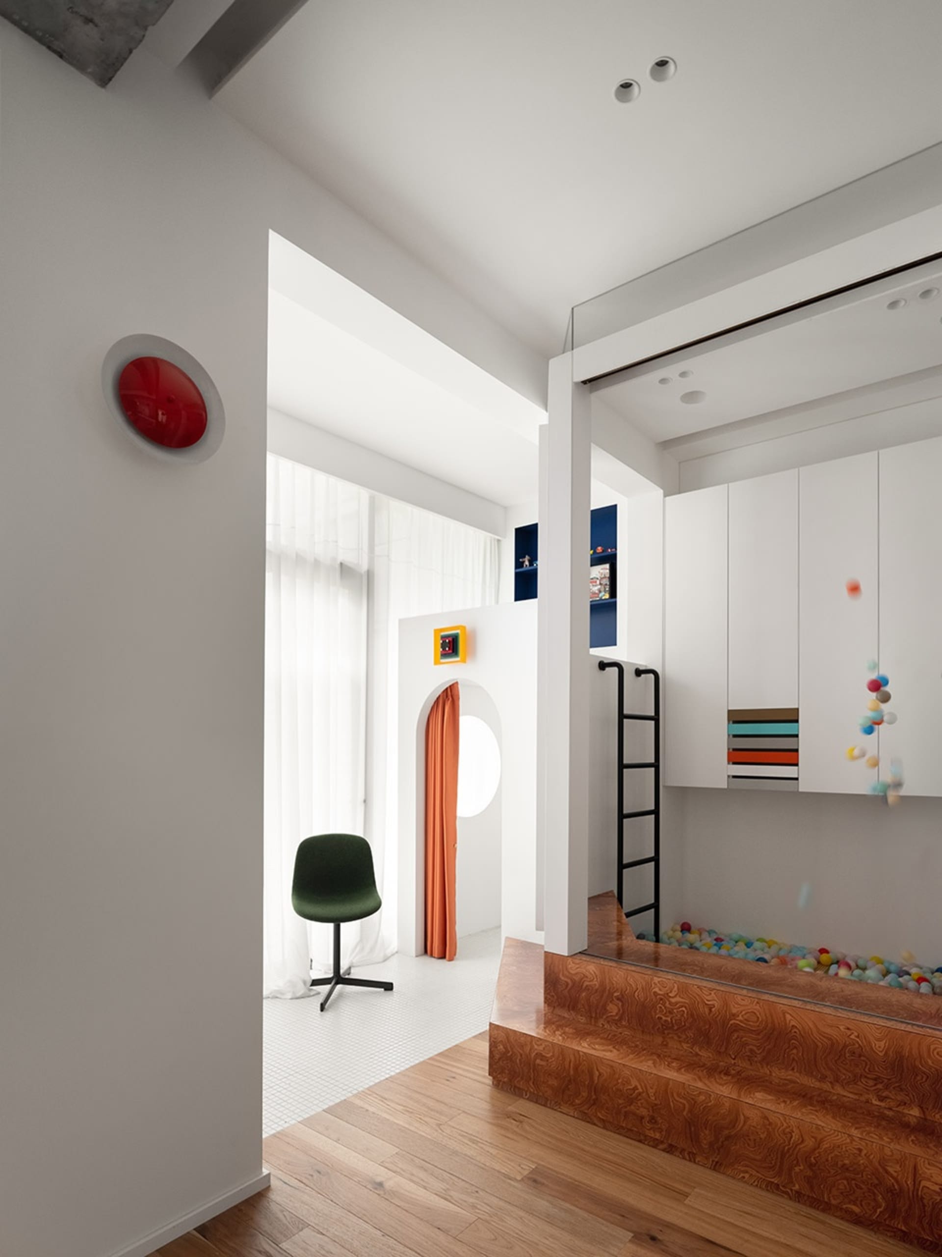 A minimalist kids playarea with a wooden floor, white walls, and colorful plastic balls in a shallow pool.