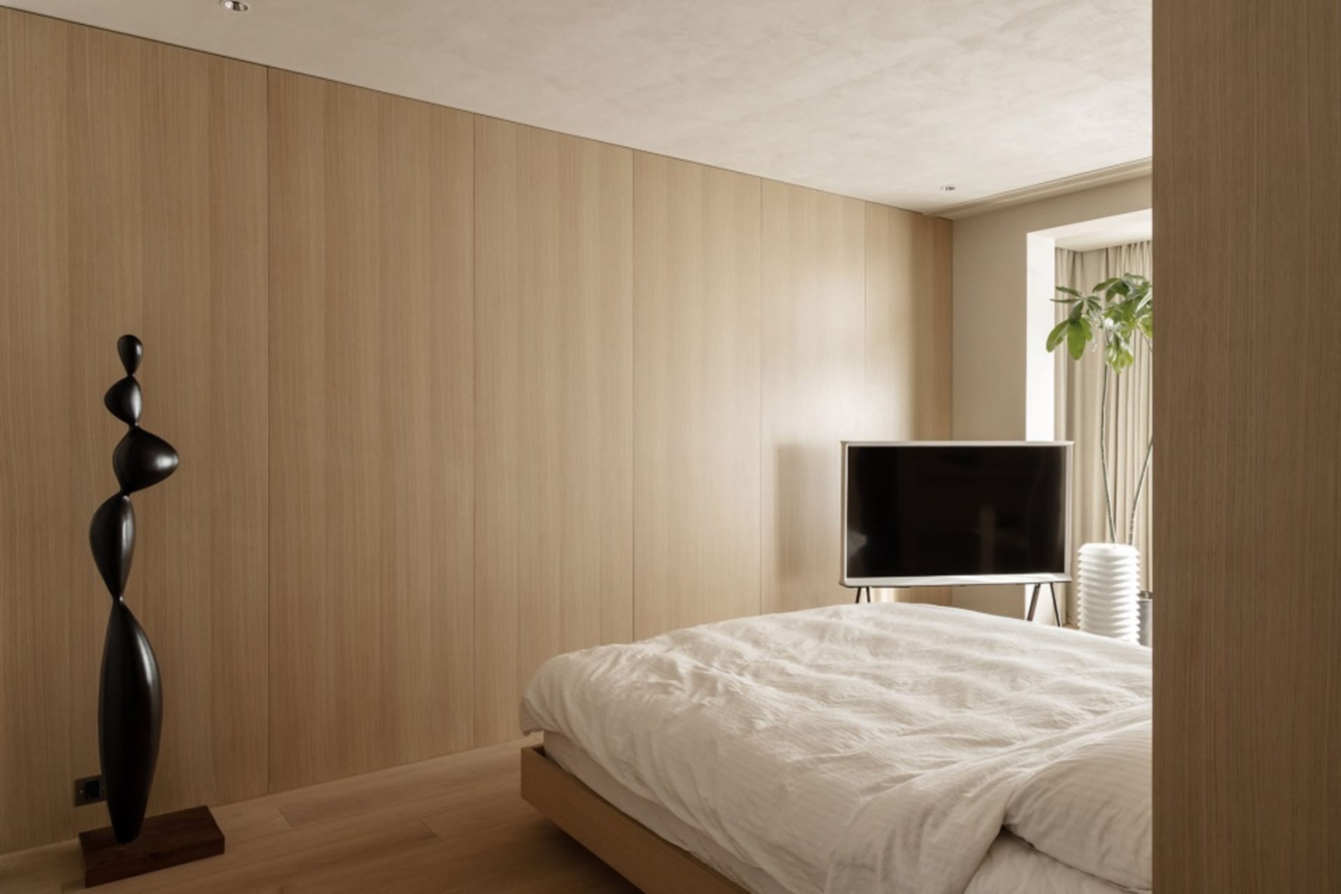 A minimalist bedroom with white wooden walls and a black abstract sculpture, featuring a bed with white sheets and a televisi