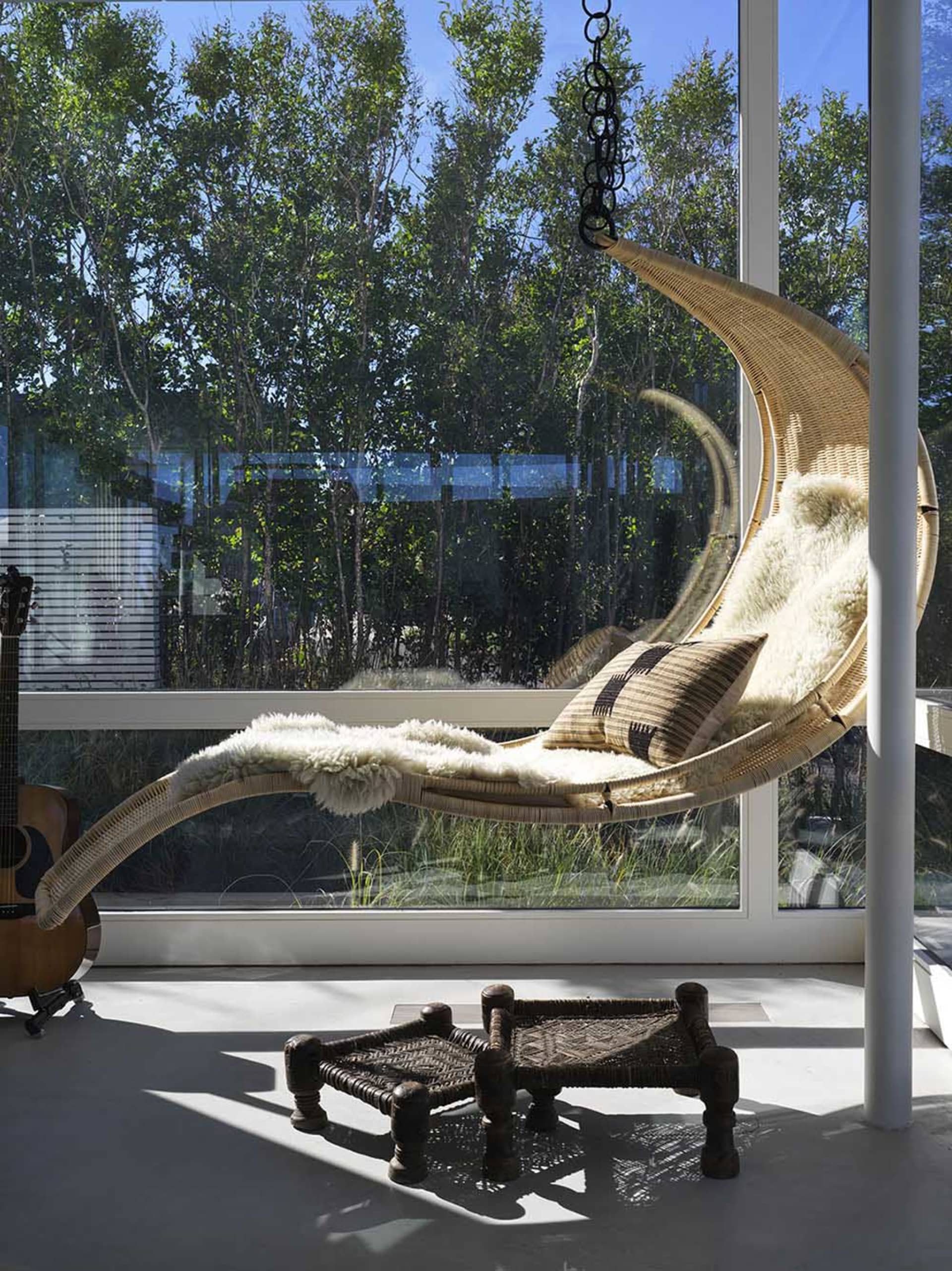 A modern indoor swing with a wicker frame and plush cushions hanging from a ceiling hook, surrounded by large windows with a