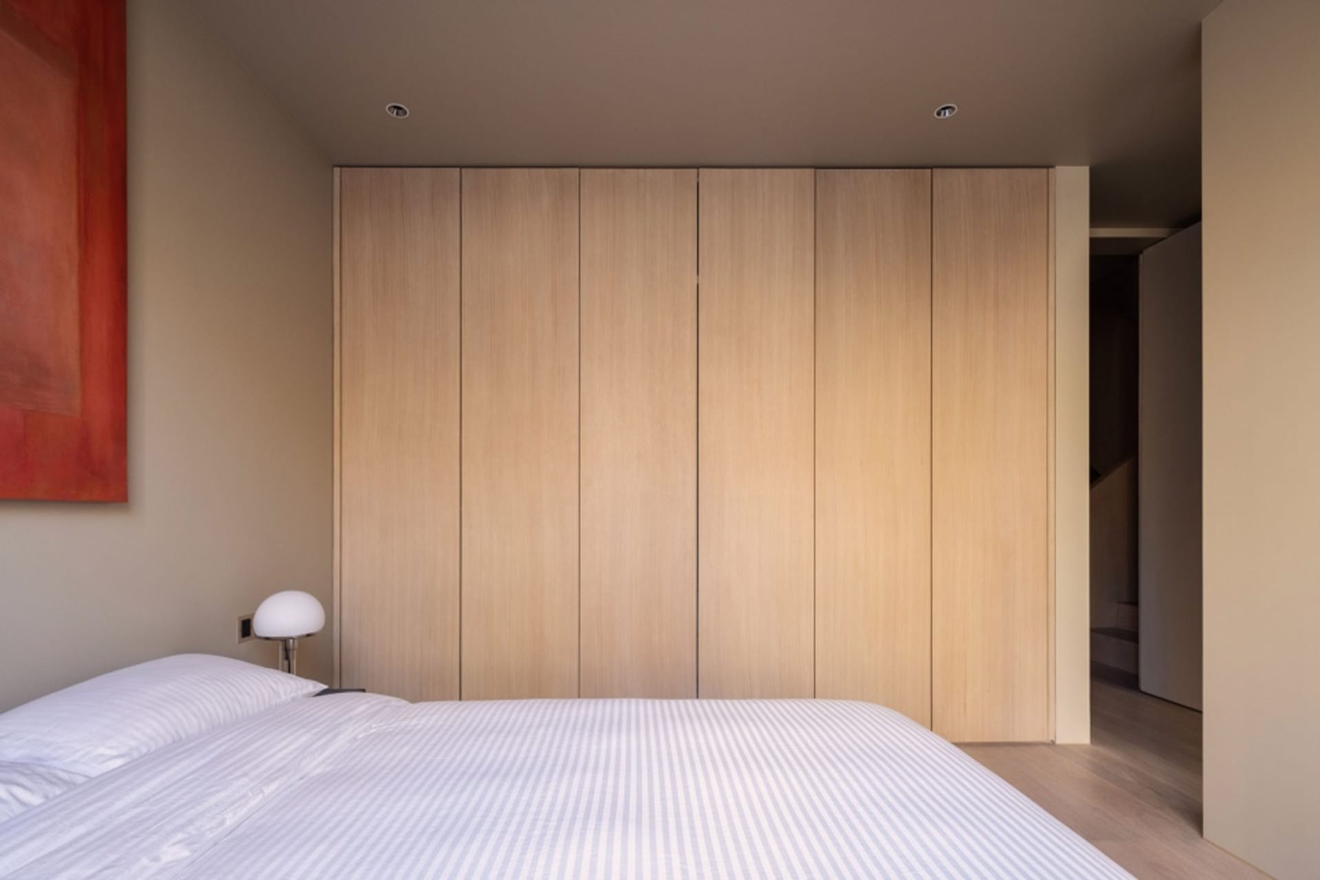 A minimalist bedroom featuring a neatly made bed with a striped white cover, a modern lampshade, and a wooden paneled wall.