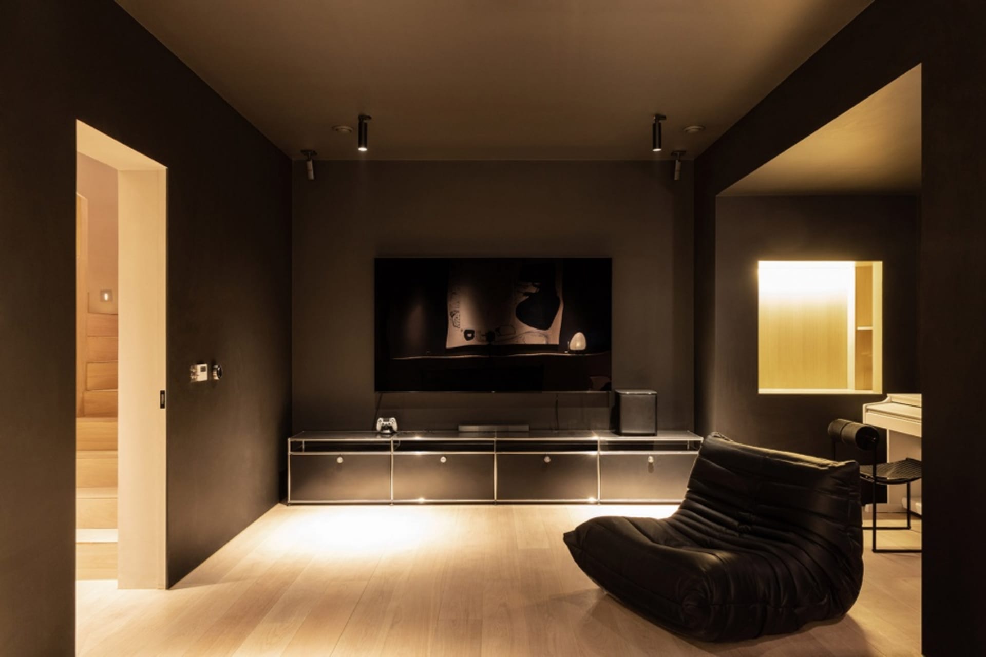A modern minimalist living room with a black chair, wooden floor, and a television mounted on a black wall.