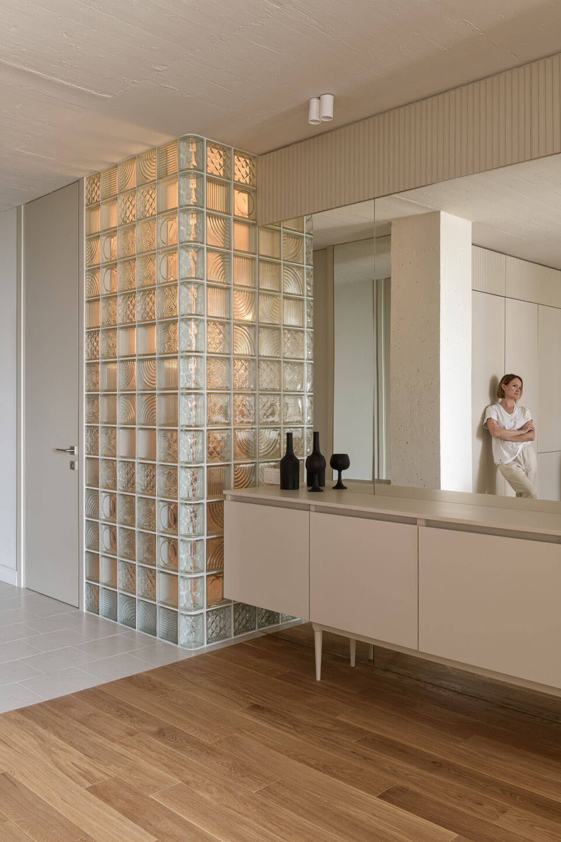A modern interior featuring glass blocks and concrete furniture, with a woman leaning against a wall.