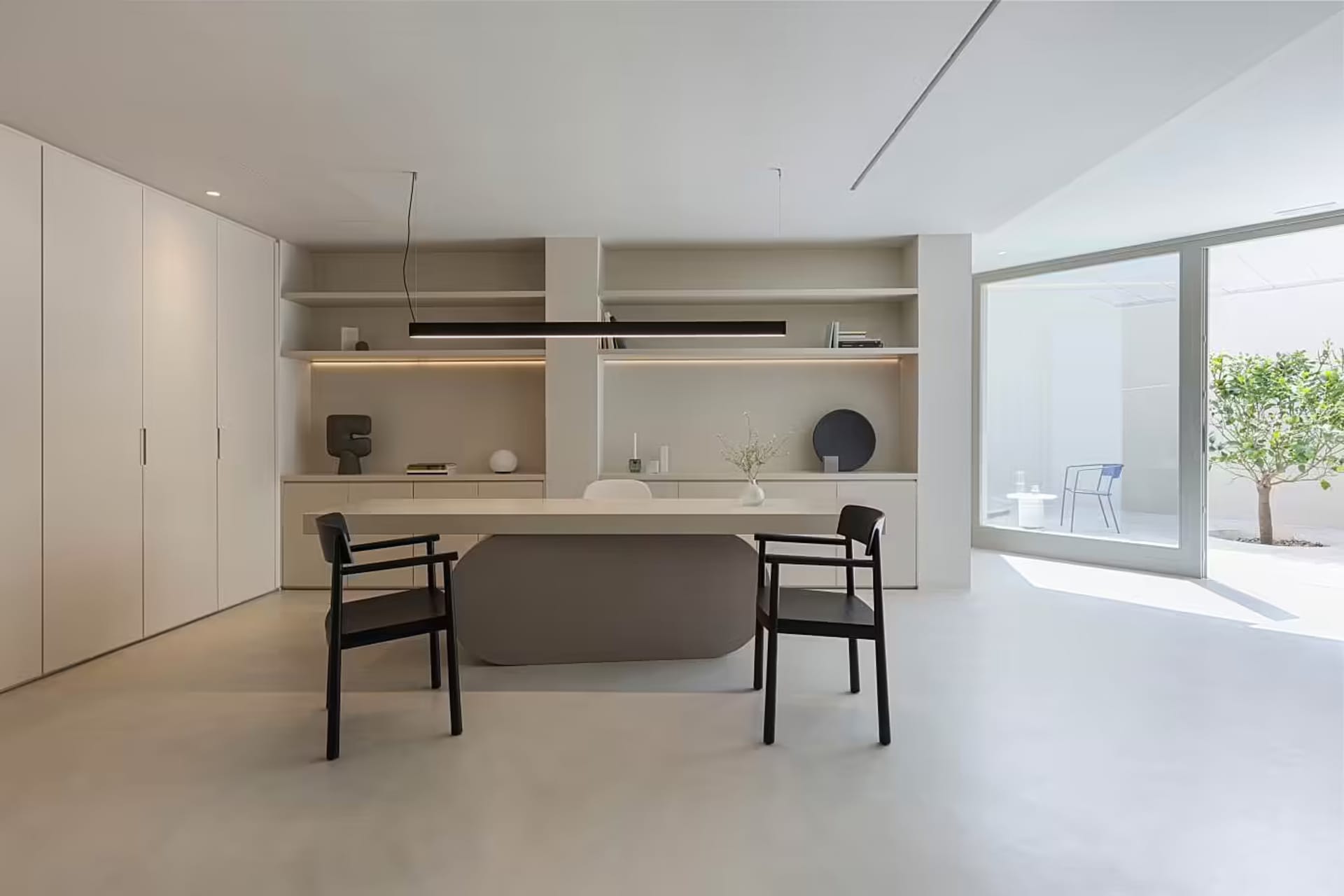 A modern minimalist office with a concrete desk, black chairs, and a sleek, white storage unit with built-in shelves.