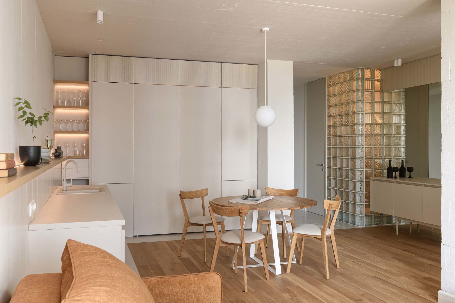Modern minimalist kitchen featuring a round wooden table, wooden chairs, and white cabinets with glass block wall.
