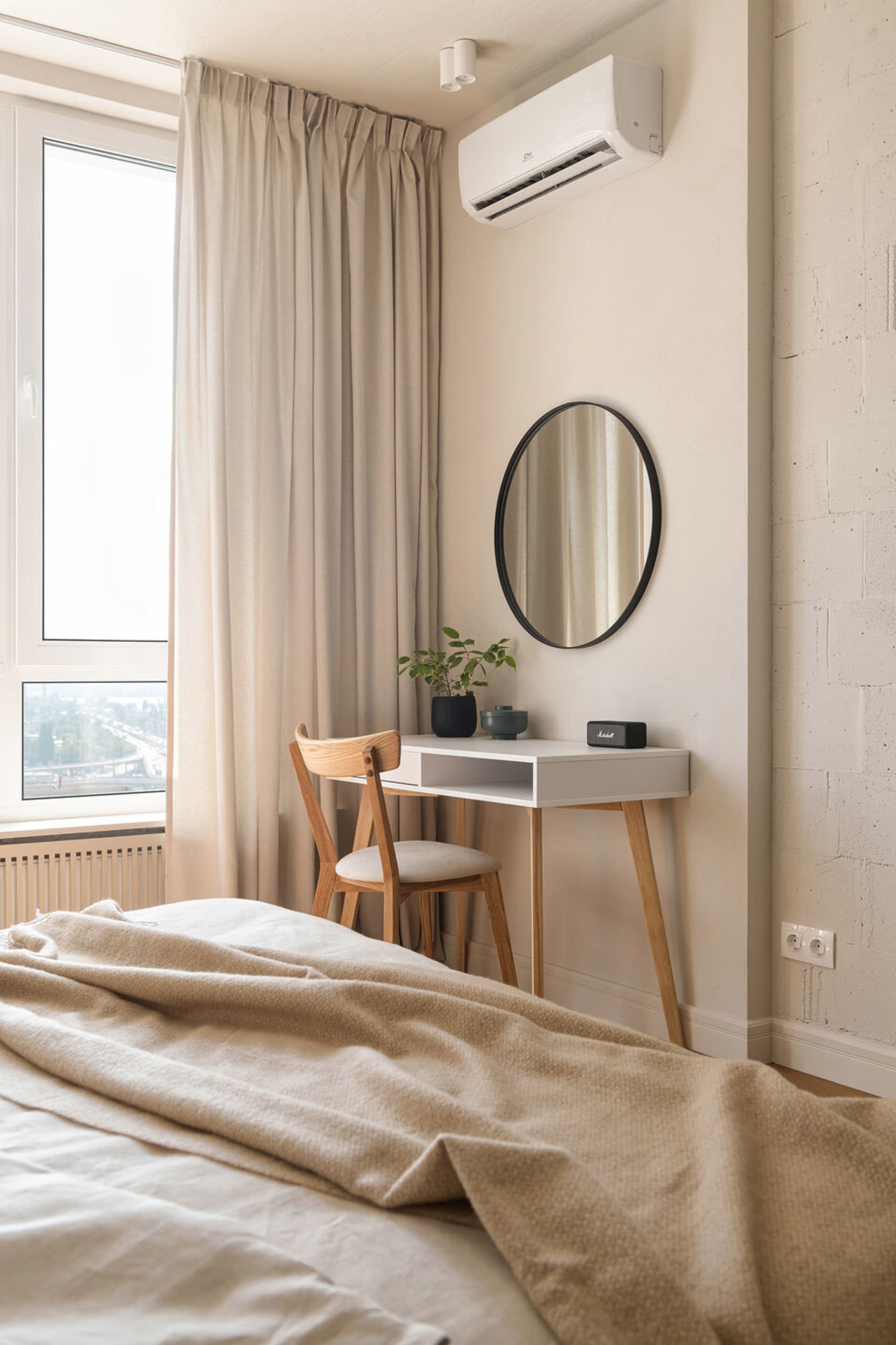 A minimalist bedroom with neutral tones, featuring a bed with beige covers, a desk with a circular mirror, and beige curtains