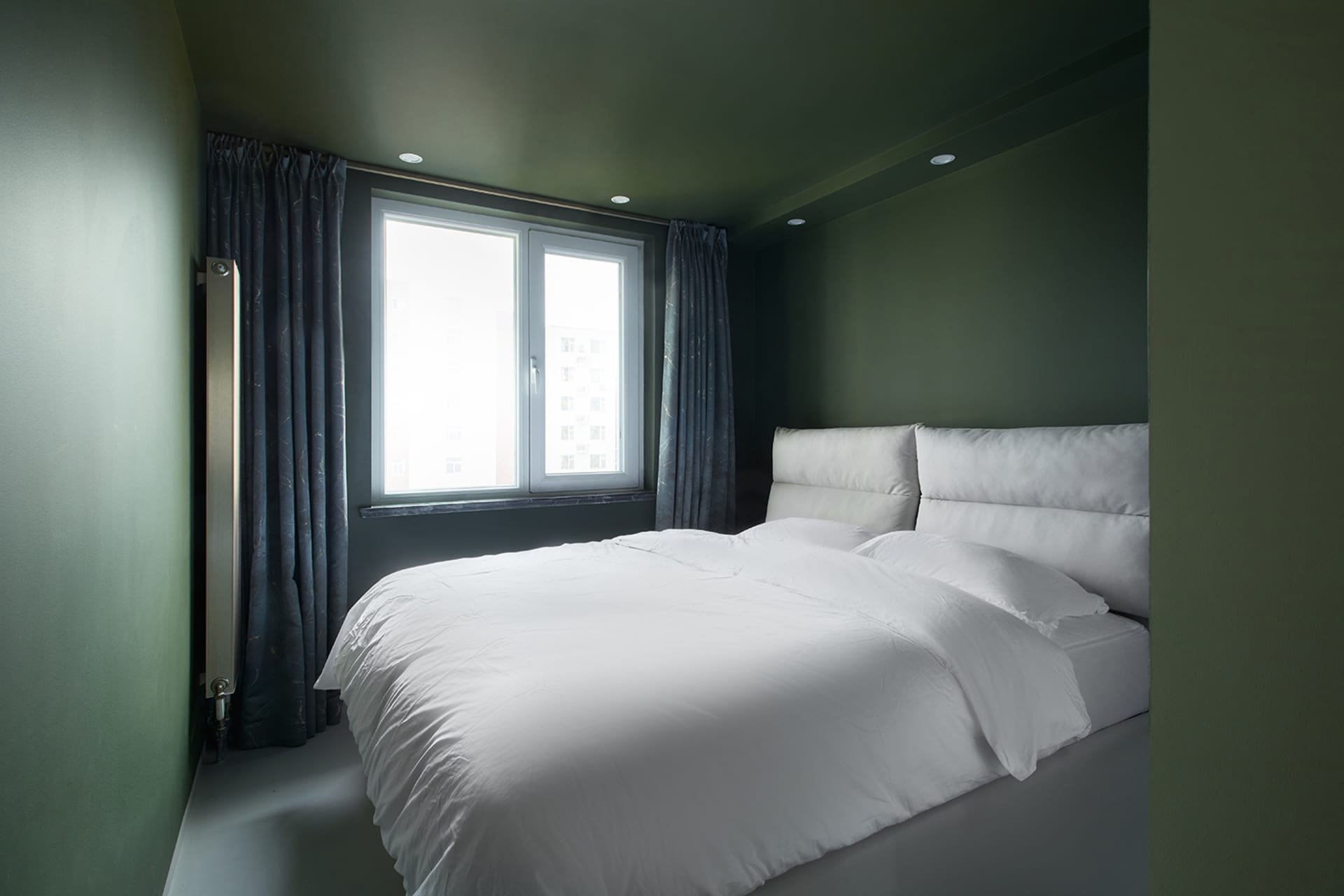 A clean, minimalist bedroom with a large bed, white bedding, and natural light coming through the window.