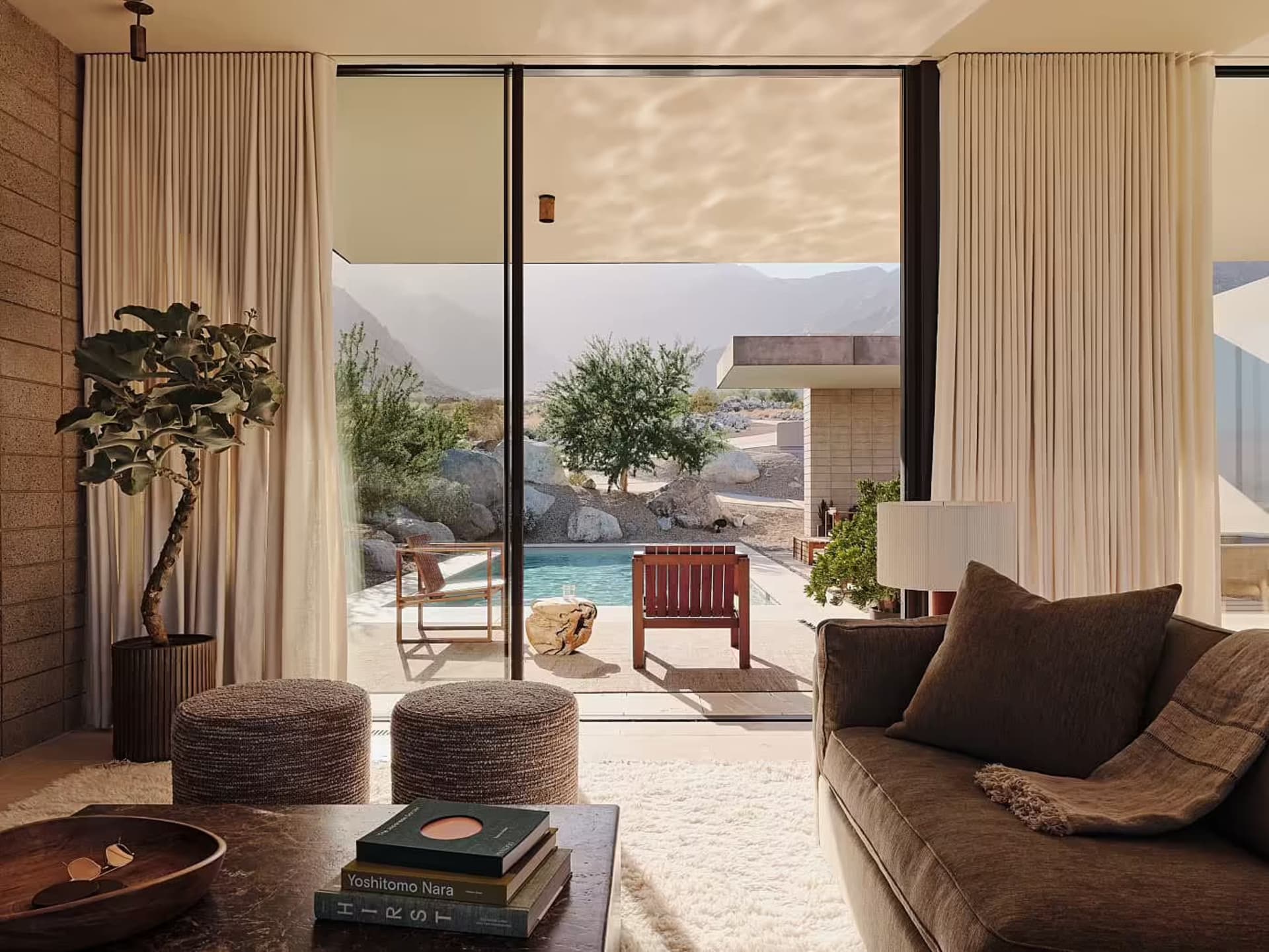 A modern minimalist living room with a large window, a comfortable sofa, and a view of a pool and mountains.