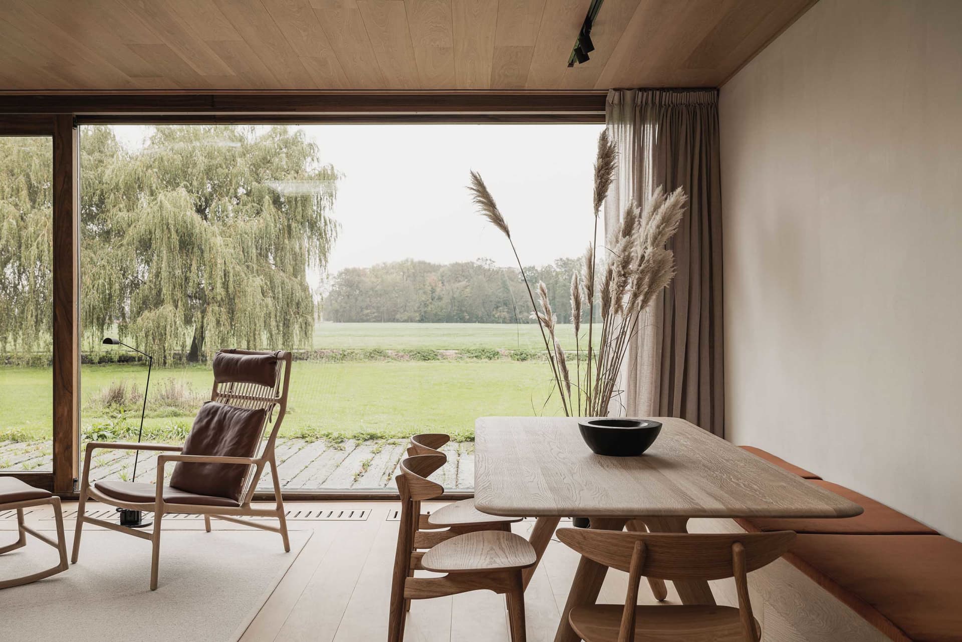 Modern living room featuring wooden furniture, natural light, and a view of green fields through large windows.