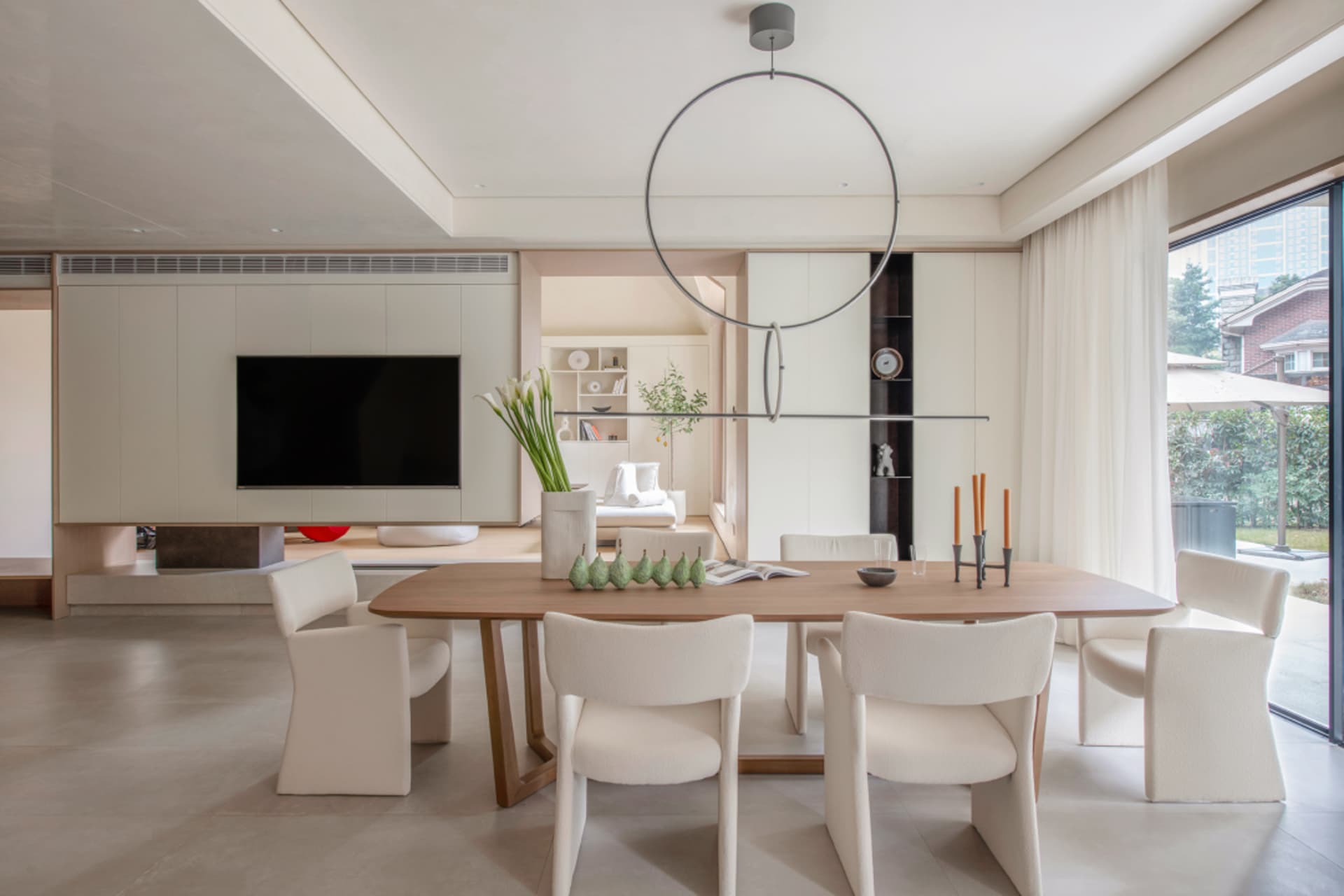 Modern minimalist dining room with white chairs and a wooden table, featuring a round hanging light fixture and a TV in the b