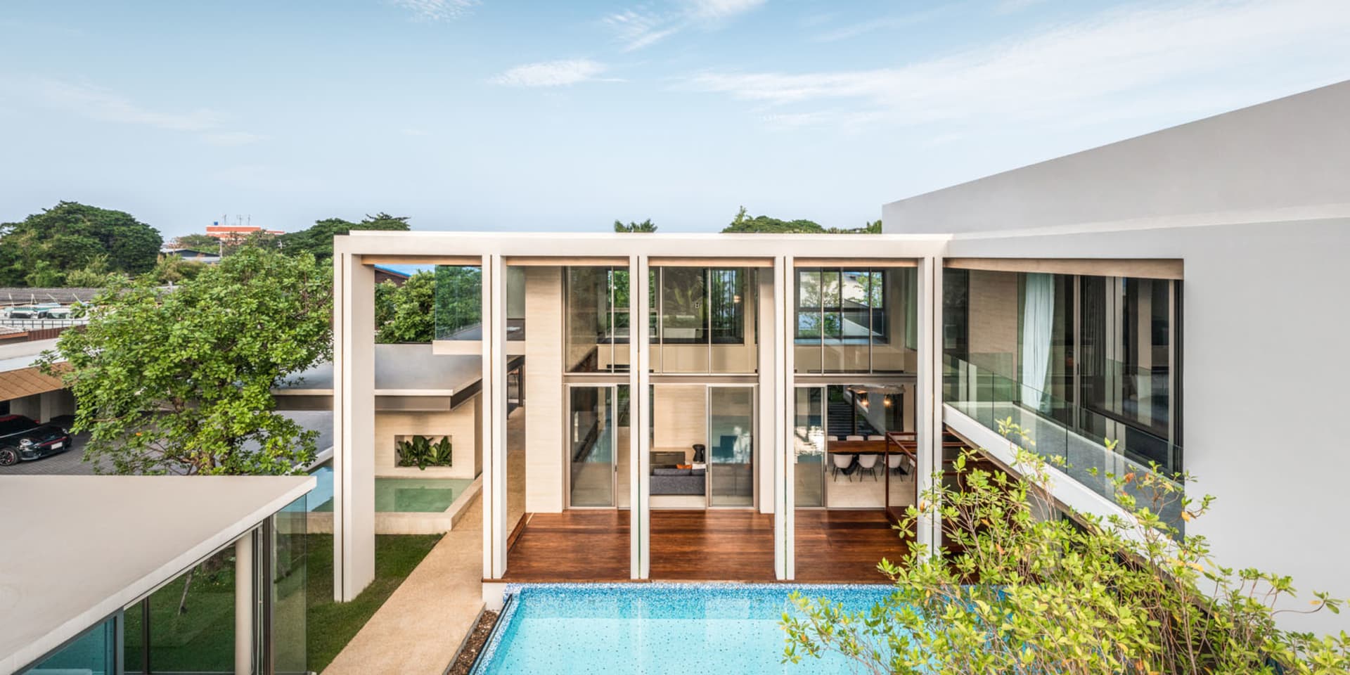 A modern residential house with glass walls and wooden flooring, featuring a swimming pool surrounded by greenery.
