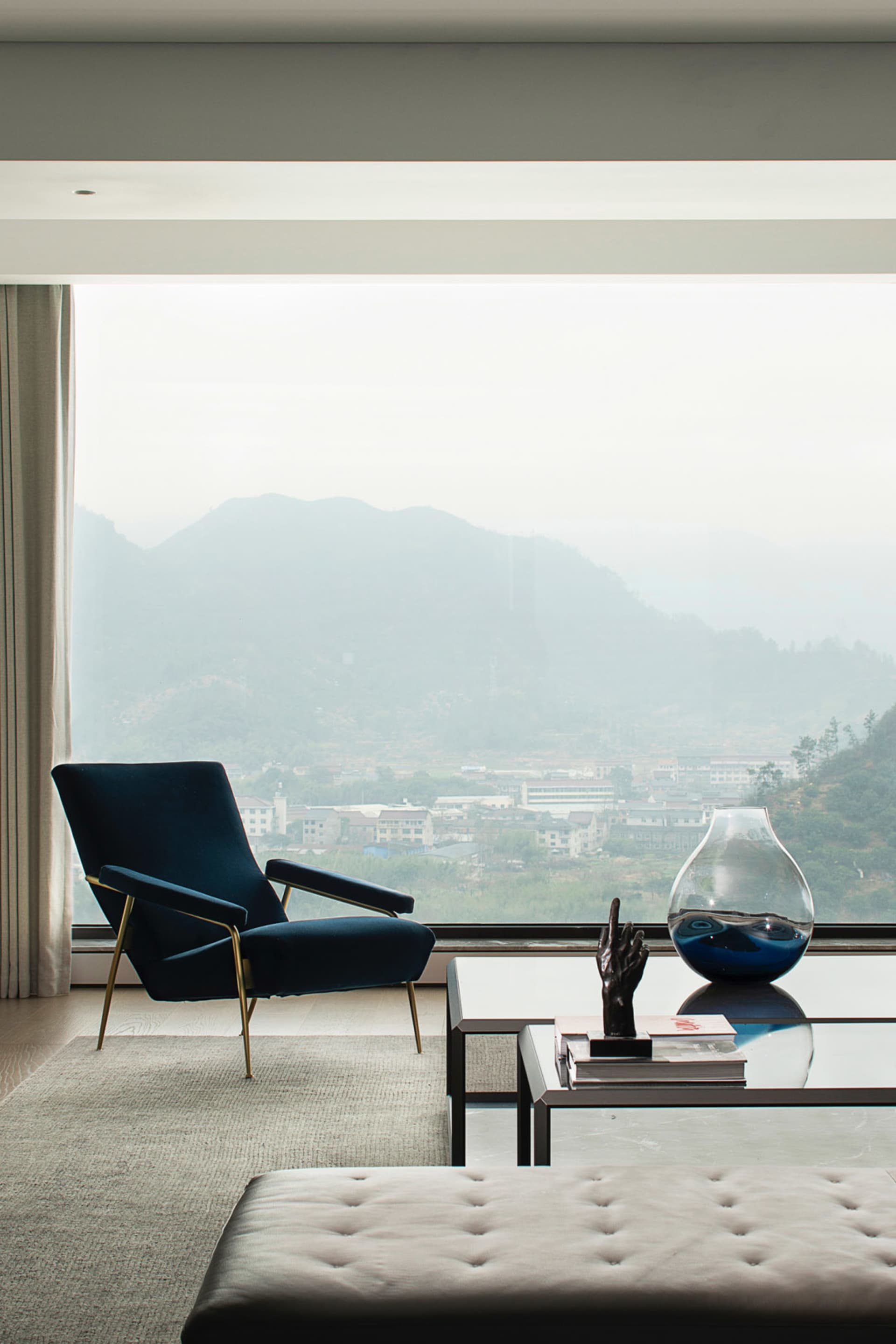 Modern living room featuring a glass coffee table, plush carpet, and a velvet armchair by a large window.