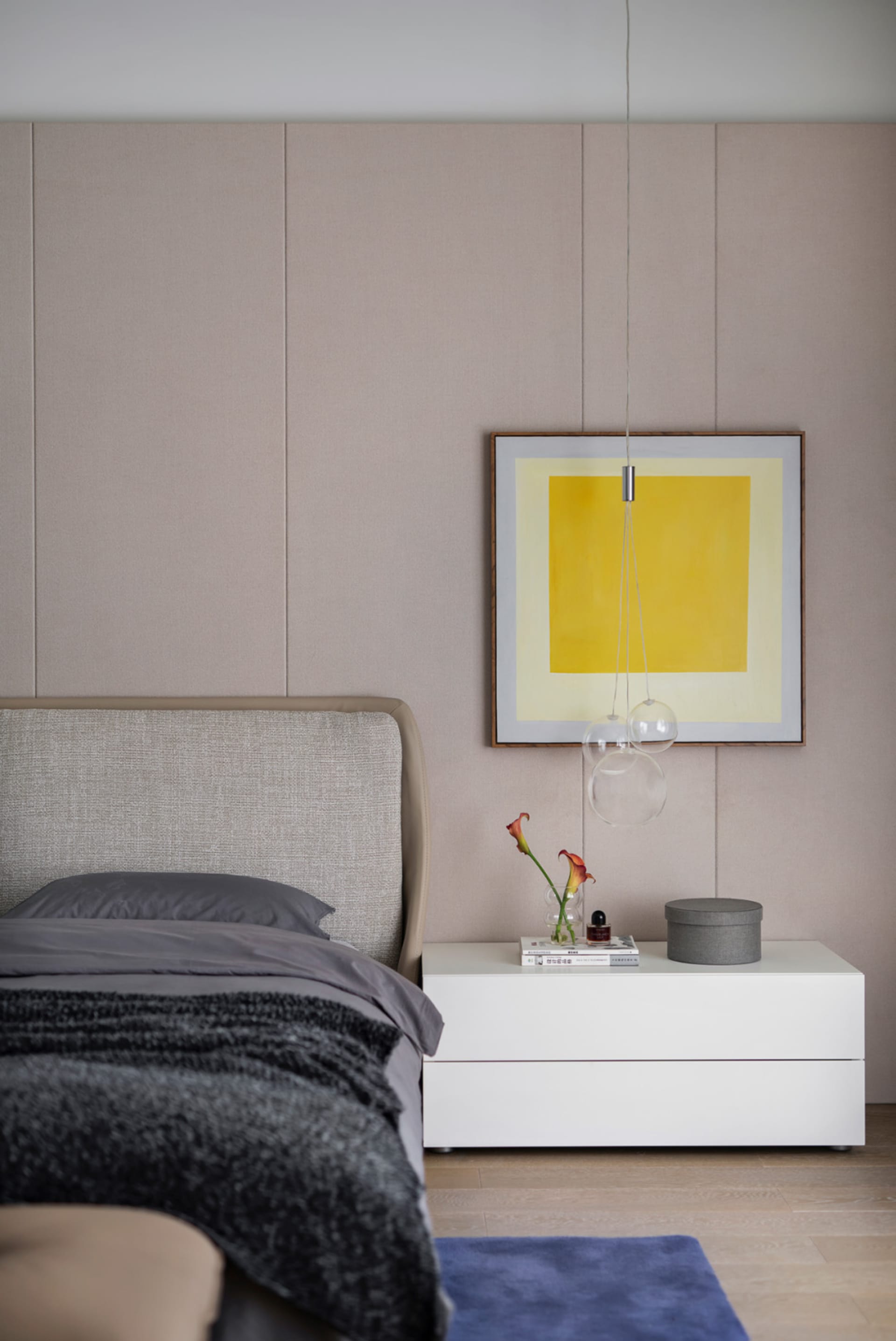 Modern minimalist bedroom featuring a fabric upholstered bed, wooden nightstand, glass vase with flowers, and abstract art.