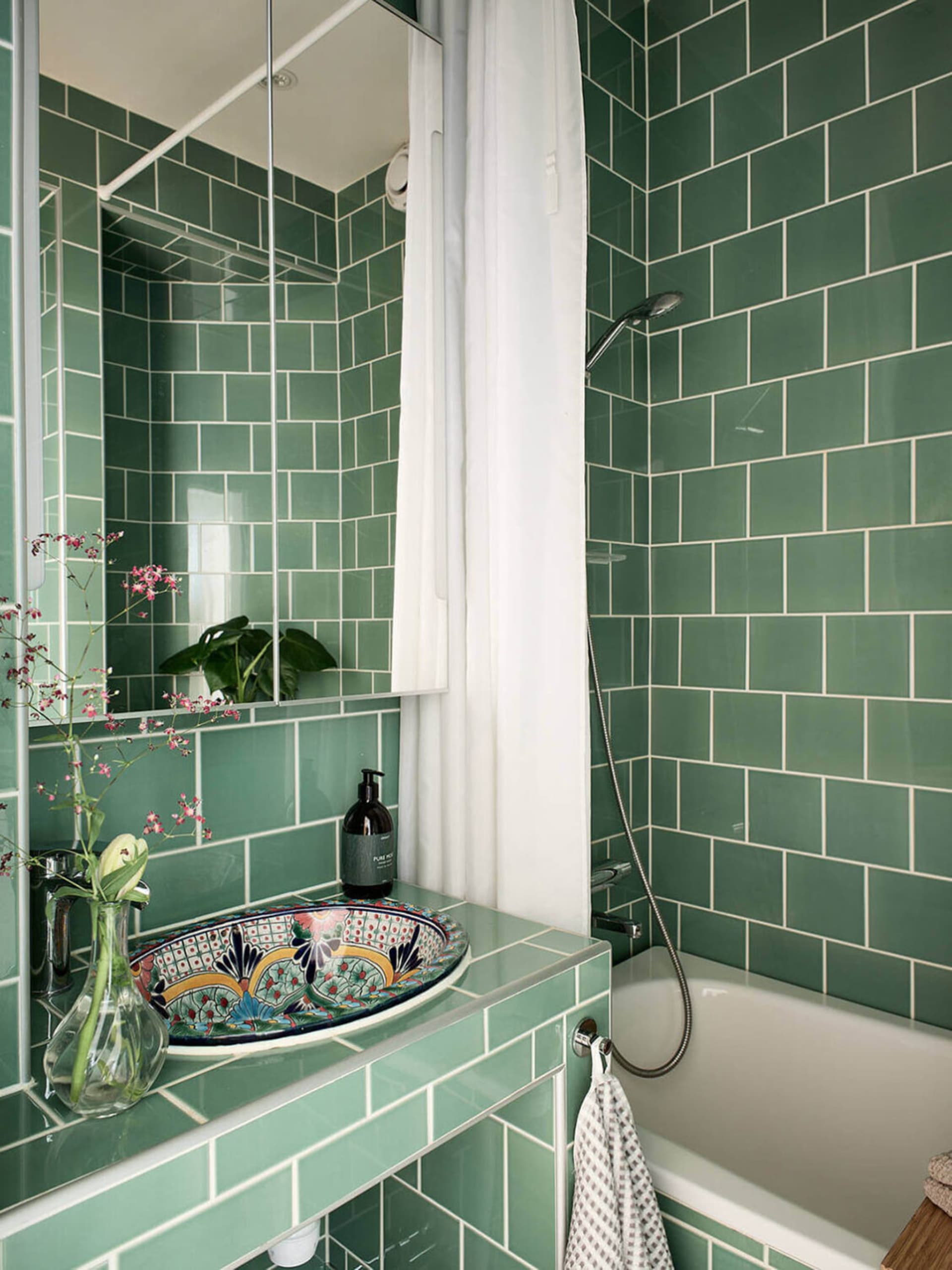 A modern bathroom with green ceramic tiles, a colorful sink, and a white bathtub.
