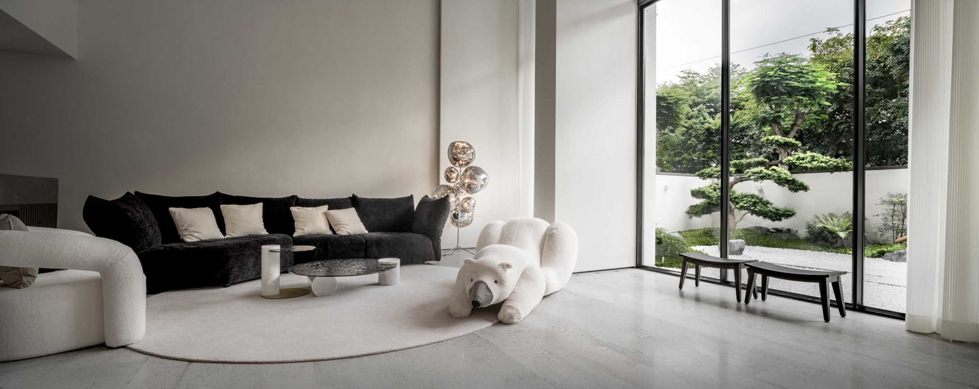 Modern minimalist living room with white marble floor, black sofa, and large windows overlooking greenery.