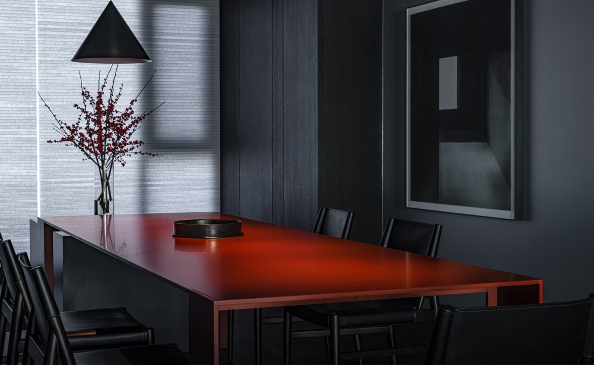 A minimalist dining room features a red tabletop with a vase of red berries and a black pendant light.