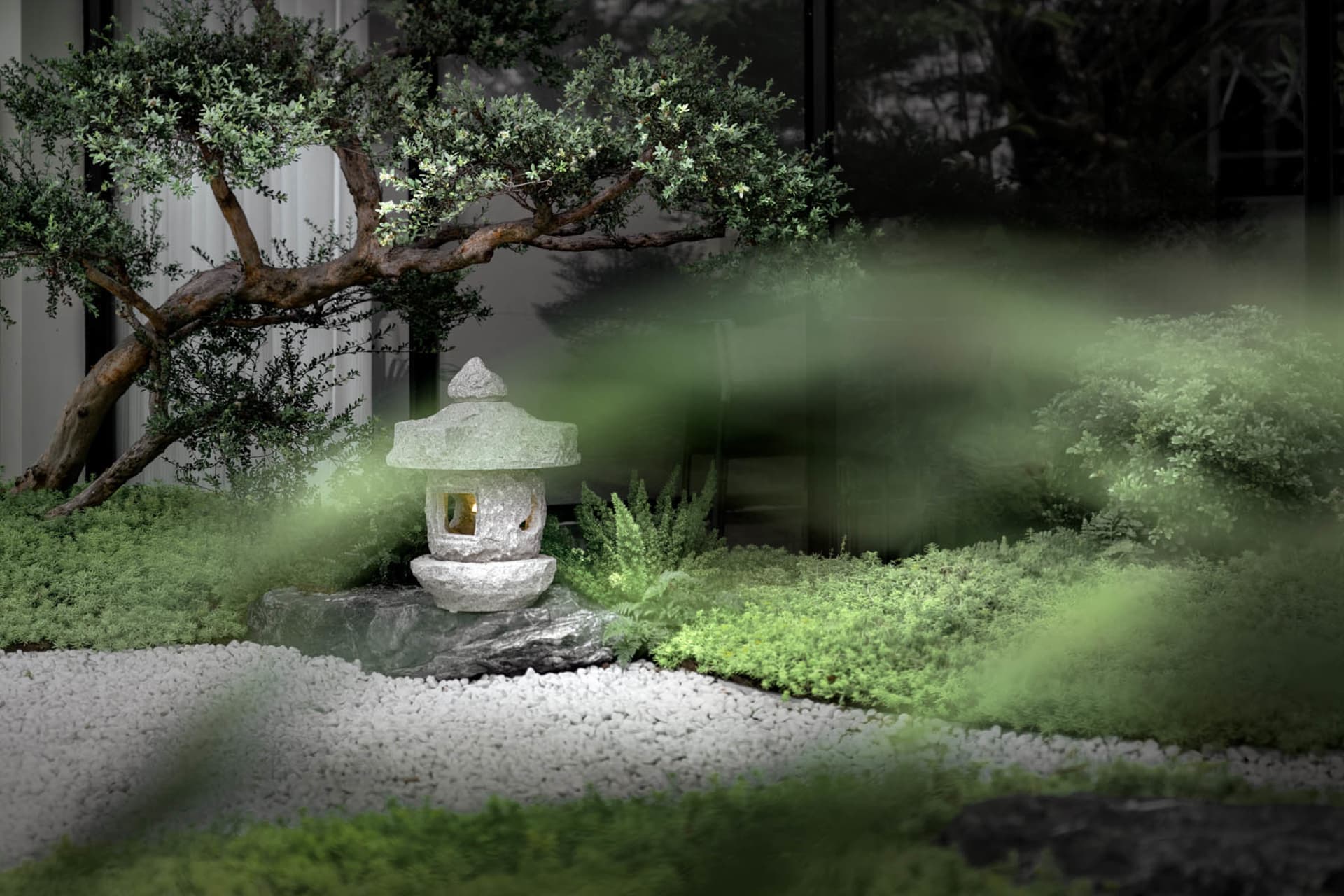 A minimalist rock garden featuring a stone lantern and a small tree, surrounded by green plants and white gravel.