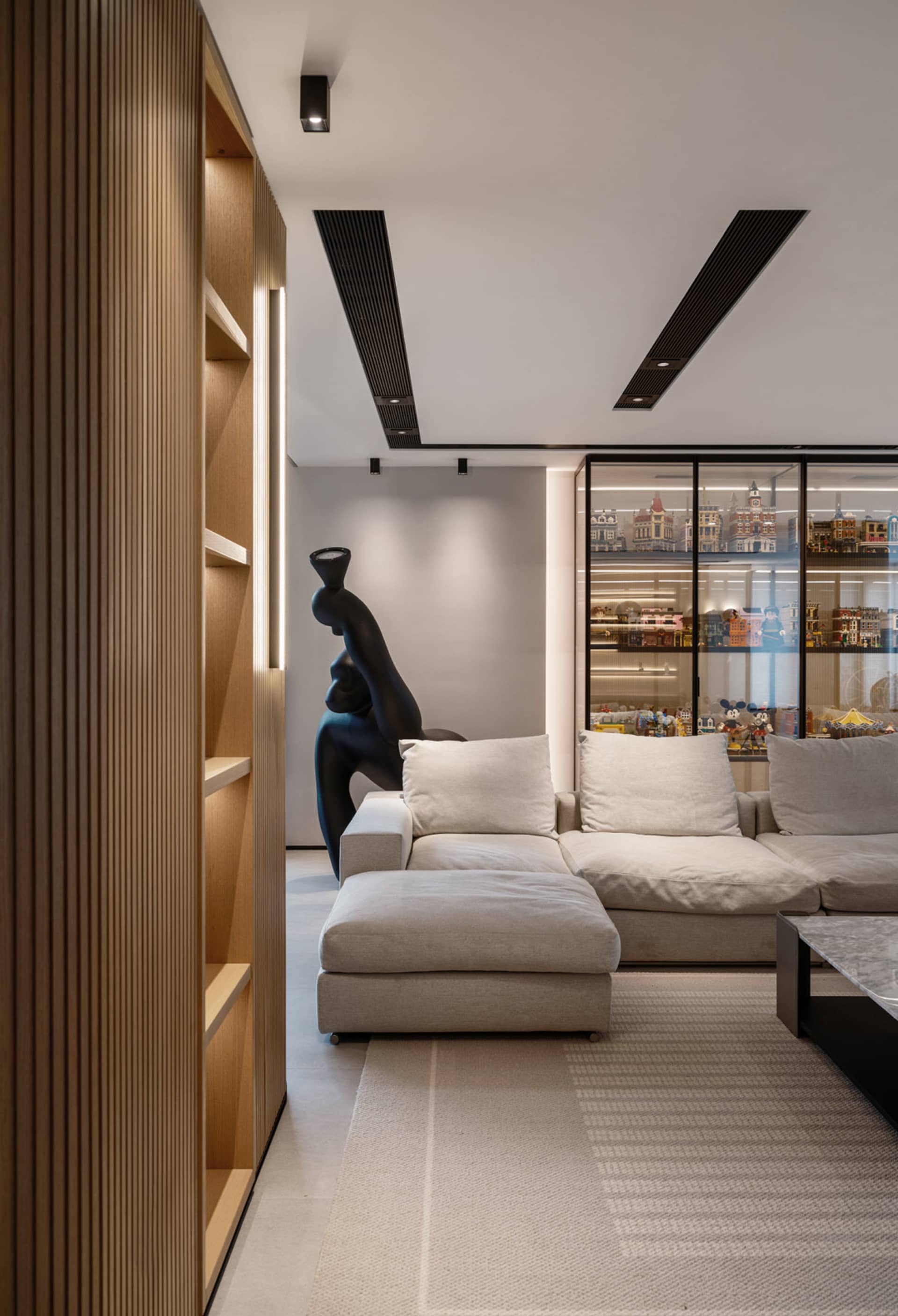 A modern, minimalist living room with a beige sectional sofa, a black sculpture, and a glass cabinet displaying various objec