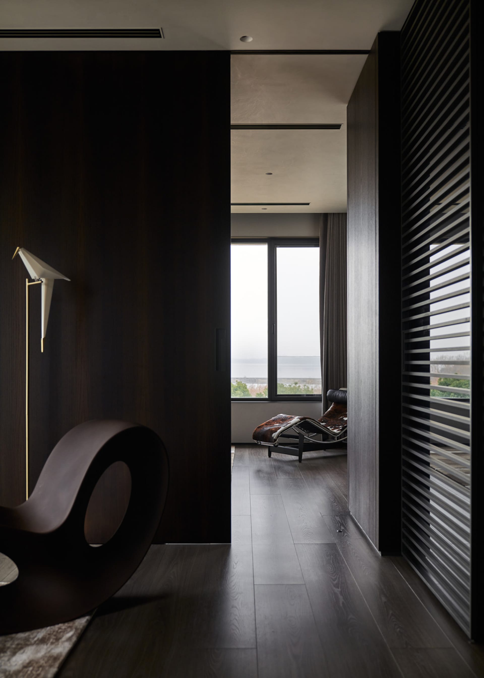 A minimalist residential interior featuring dark wooden floors, a sleek, modern armchair, and a lounge chair with a leather u