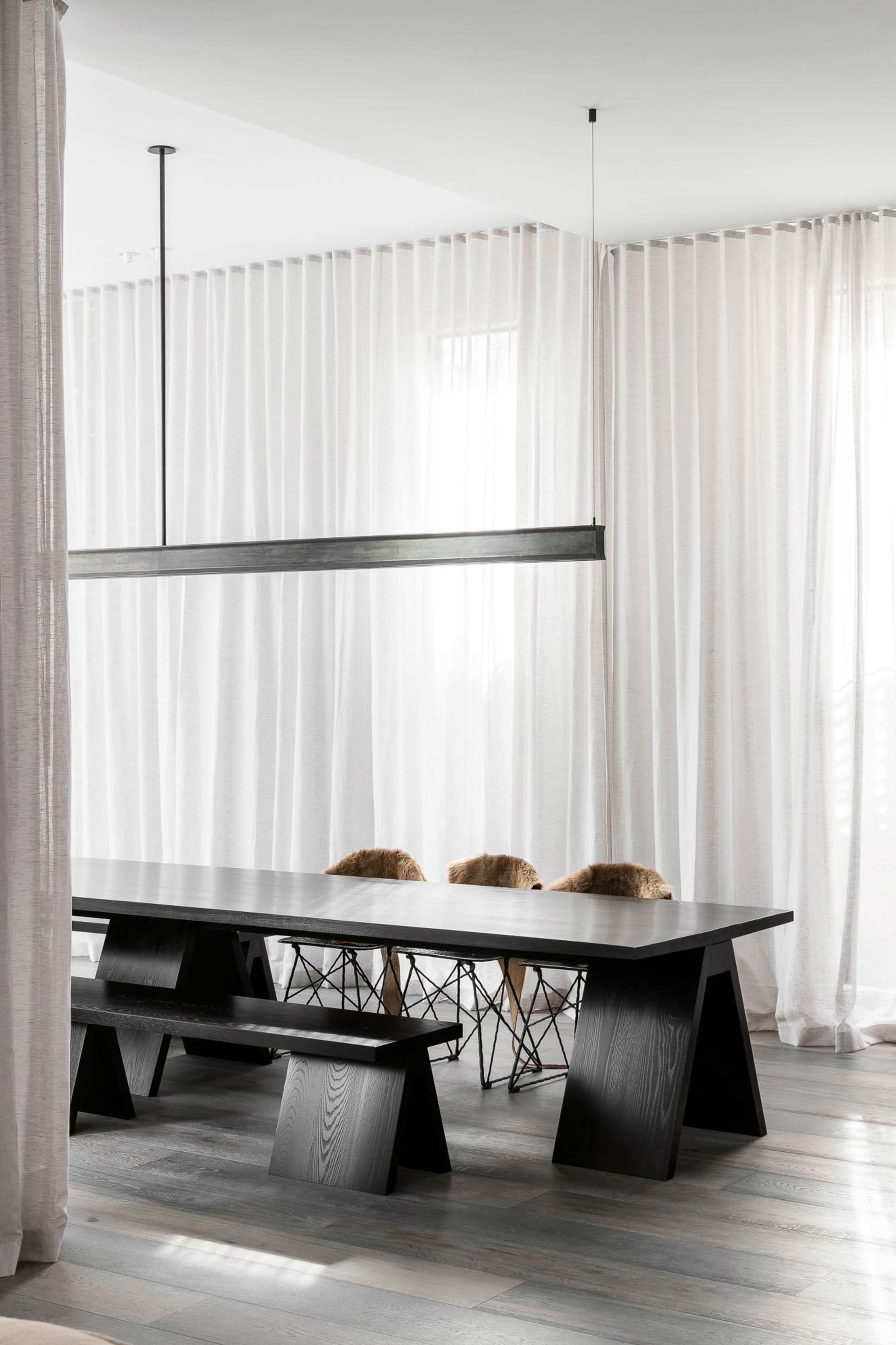 A minimalist dining room with a black table, white curtains, and wooden flooring.